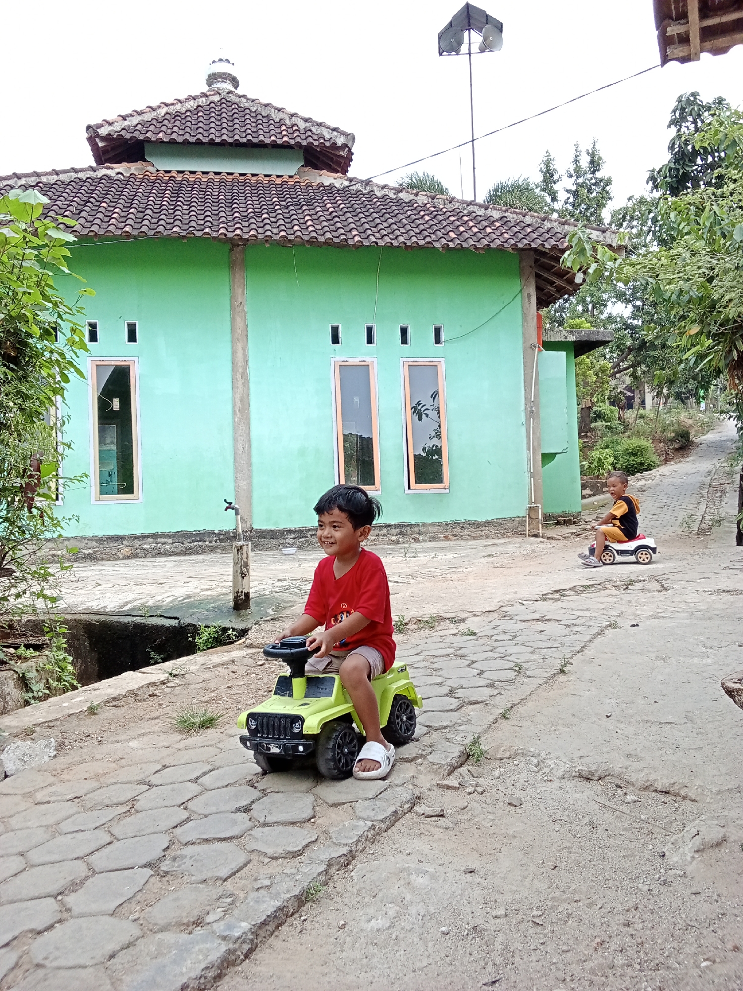 In Pringsewu Barat, Indonesia, two young boys are playing outside. A boy in a red shirt and shorts is seated on a green toy jeep, steering it. He is positioned on a paved path that leads away from a light green building with a dark tiled roof. Another boy, wearing a yellow shirt and shorts, is riding a white toy car further down the path. The building appears to be a mosque, evidenced by the dome on its roof and the speaker system mounted on a pole in front of it. Lush green foliage surrounds the scene, with trees and plants visible on the right and left sides of the frame. The sky is overcast.