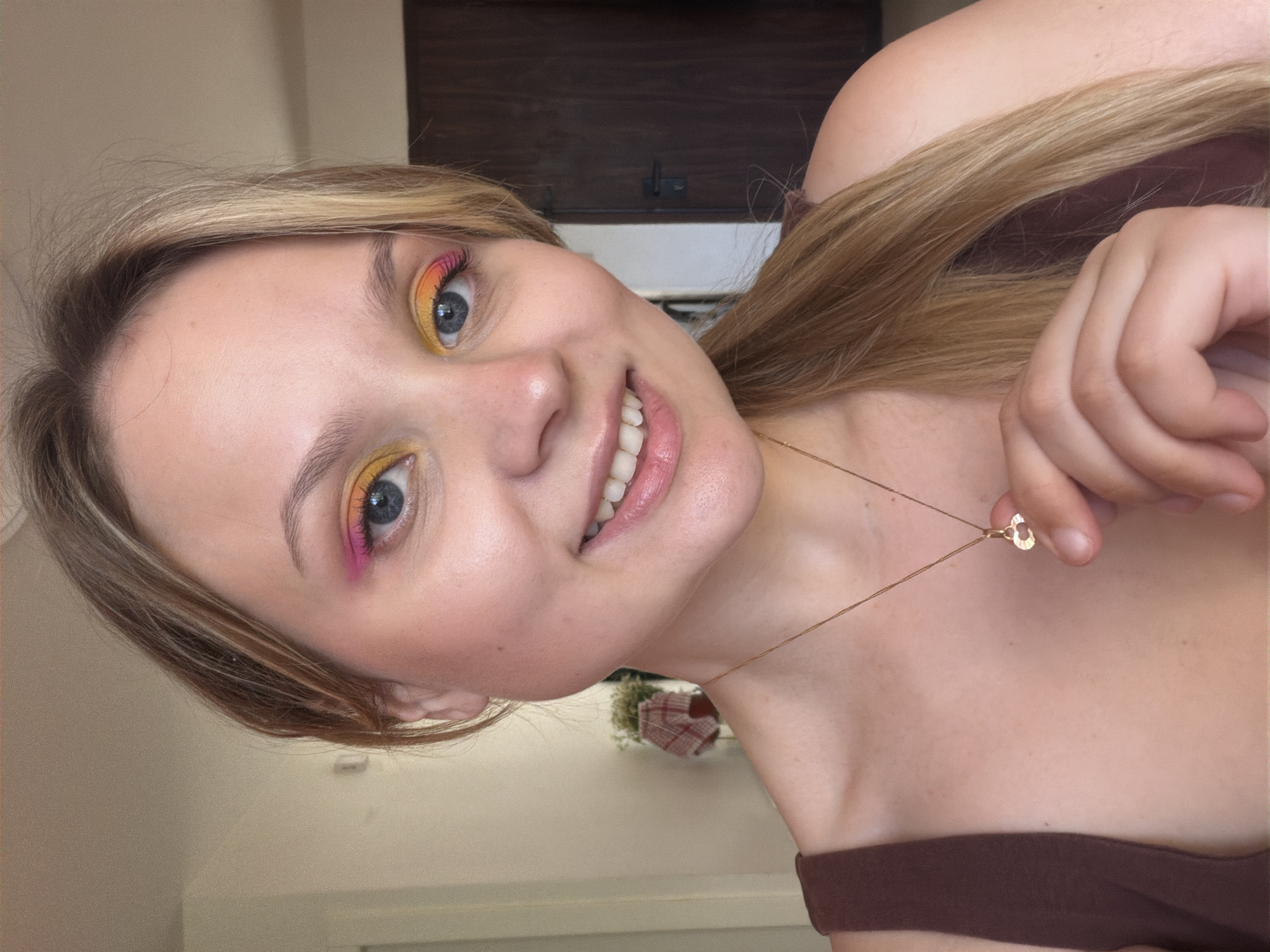A young woman with blonde hair smiles at the camera. She is wearing a brown top and a gold necklace. She has a colorful eyeshadow look, with pink, orange, and yellow shades. The background is blurry and shows a dark wooden cabinet and a white door. The location is not discernible from the image.