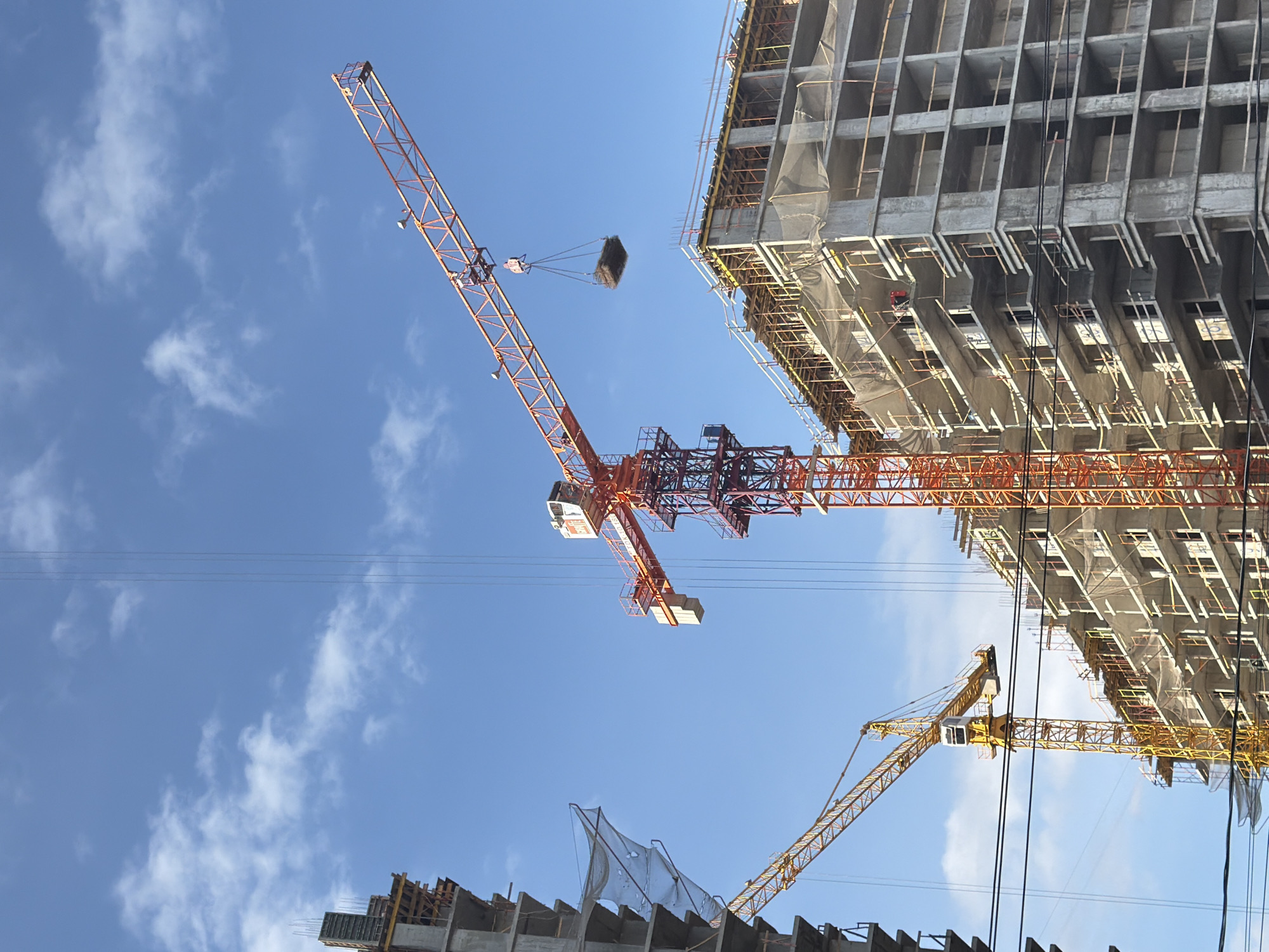The image shows a construction site in Rostov-on-Don, Russia. Two cranes are visible. An orange crane is lifting a load toward a concrete building under construction. Another yellow crane is in the lower right corner of the image. Power lines run across the blue sky. The unfinished building has scaffolding and protective netting. Another building under construction is partially visible in the lower left corner.