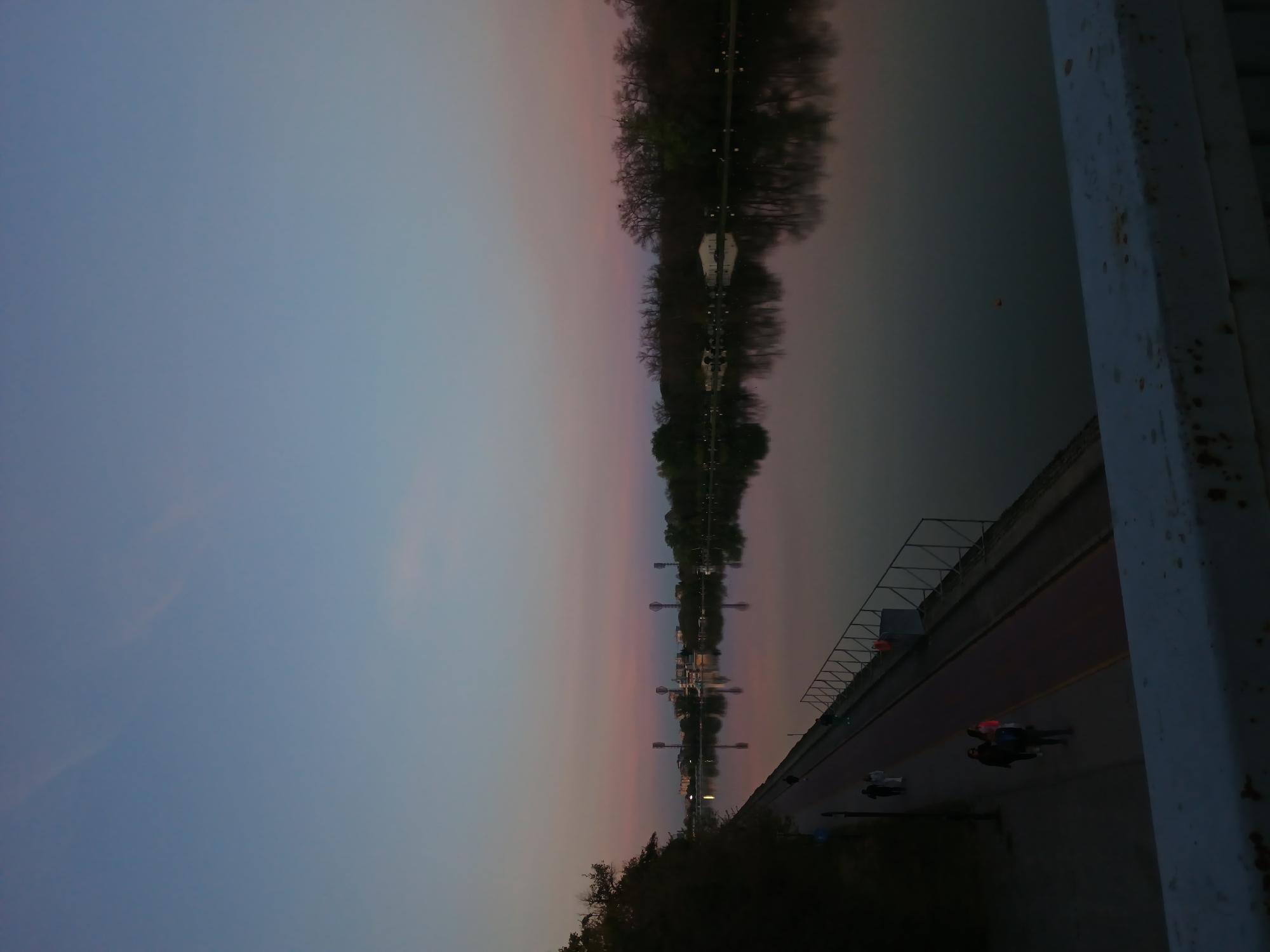 The image, initially rotated 90 degrees clockwise, presents a serene riverine landscape captured at either dusk or dawn. The sky, occupying the upper left quadrant, transitions from a soft blue to delicate pink and orange tones at the horizon line, indicative of the early or late hours of the day.
The central feature is a calm river, likely the Maritsa River in Plovdiv, Bulgaria, which perfectly mirrors the sky's gradients and the silhouetted trees lining its distant bank. These trees, appearing mostly bare or sparsely leaved, suggest a late autumn or winter setting. Scattered lights or small structures are faintly reflected in the water along the far bank.
In the foreground, on the right side of the frame, a sturdy, light-colored metal bridge structure, marked with reddish-brown rust, provides a stark contrast. Beneath this structure lies a dark-surfaced path or road. Several individuals are present on this path: one person is seen on a bicycle or scooter, and a small group of two or three people, one distinctive in a red jacket or top, are either walking or standing.
The overall impression is one of quiet contemplation, with the soft, diffused light enhancing the tranquil mood of the scene. There is no discernible text visible in the image.