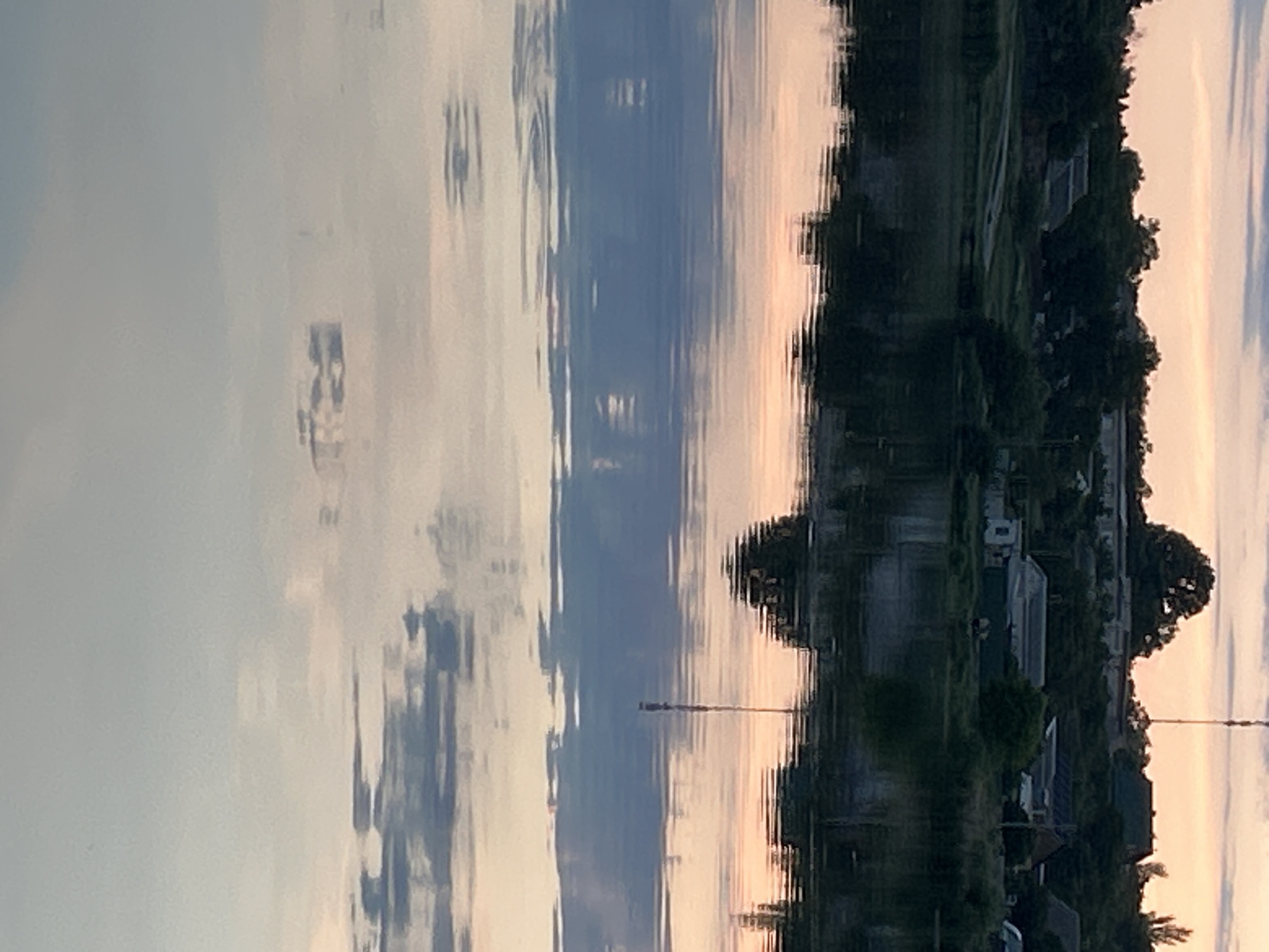 This image depicts a tranquil water scene with a village nestled on the opposite bank and a reflection of the sky in the water. The location is identified as Hnarovske, Ukraine. The water surface is calm, broken only by gentle ripples that distort the reflection of the sky and the shoreline.
On the bank, a cluster of buildings, mostly residential with pitched roofs, are visible amidst dense greenery. Trees and shrubs form a natural barrier along the water's edge. The sky above is painted with the soft hues of either sunrise or sunset, featuring light pinks, oranges, and blues. A tall communication tower rises above the tree line in the distance. The reflection in the water mirrors these elements, with the sky's colors creating a soft, diffused palette on the water's surface. The scene is devoid of any people or visible activity, suggesting a peaceful and undisturbed moment.