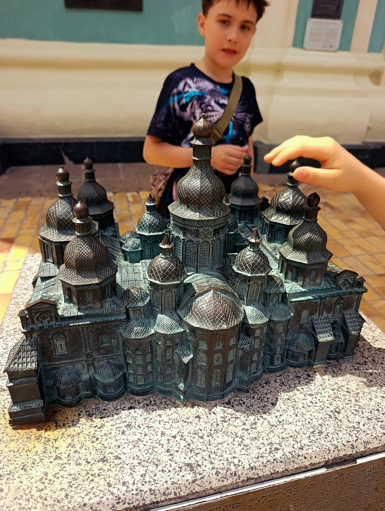 In Kyiv, Ukraine, a boy stands behind a detailed miniature replica of St. Sophia Cathedral, which is made of bronze and rests on a granite pedestal. The boy is wearing a black t-shirt with a blue and white abstract design and a light brown strap is slung across his shoulder. Another person's hand, seemingly a child's, is reaching out towards the miniature cathedral, with their index finger hovering over one of its domes. The background shows a wall with a light teal and cream color scheme. The ground is paved with light brown tiles. The lighting suggests it is daytime, with shadows cast by the miniature cathedral.