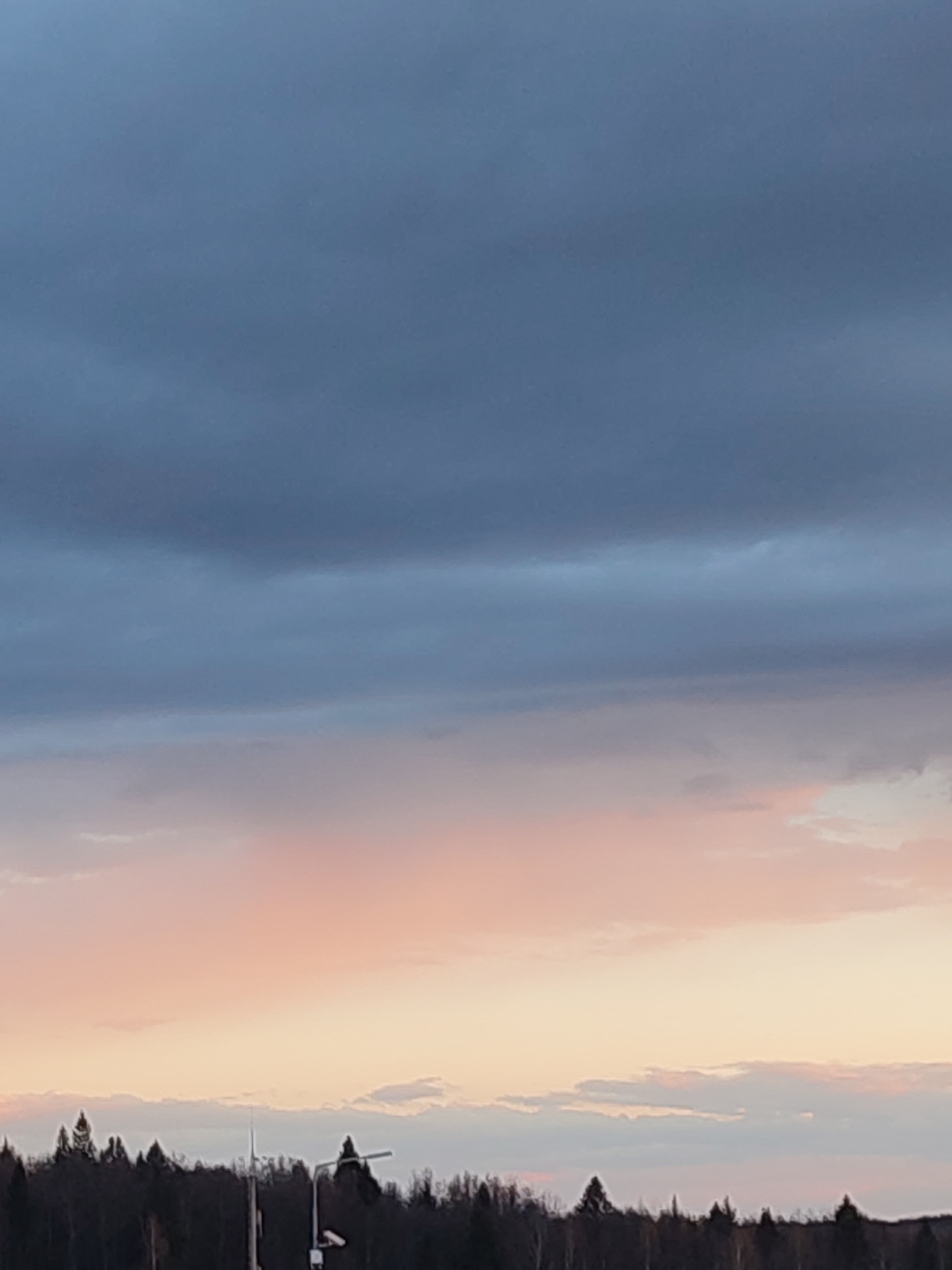 This media file captures a serene twilight landscape, dominated by a vast sky transitioning through various hues. The upper portion of the sky is a deep, muted blue-grey, suggesting either the onset of night or the pre-dawn hours. Below this, layers of lighter blue-grey clouds extend across the frame. Towards the horizon, the sky transforms into soft pastel shades of pink, orange, and light yellow, illuminated by the low sun, indicative of either sunrise or sunset. Scattered clouds, some wispy and some more defined, are visible, particularly catching the warm light near the horizon.
A dense line of dark trees forms the horizon across the bottom quarter of the image, appearing largely silhouetted against the lighter sky. These trees have the distinct conical shape of conifers. Amidst the trees, a tall, slender, light-colored pole or mast is visible slightly to the left of the center. To its right, another structure, possibly a street light with a horizontal arm, stands out.
The scene conveys a peaceful and calm atmosphere, characteristic of twilight hours in a rural or semi-rural setting. There are no visible people or text.