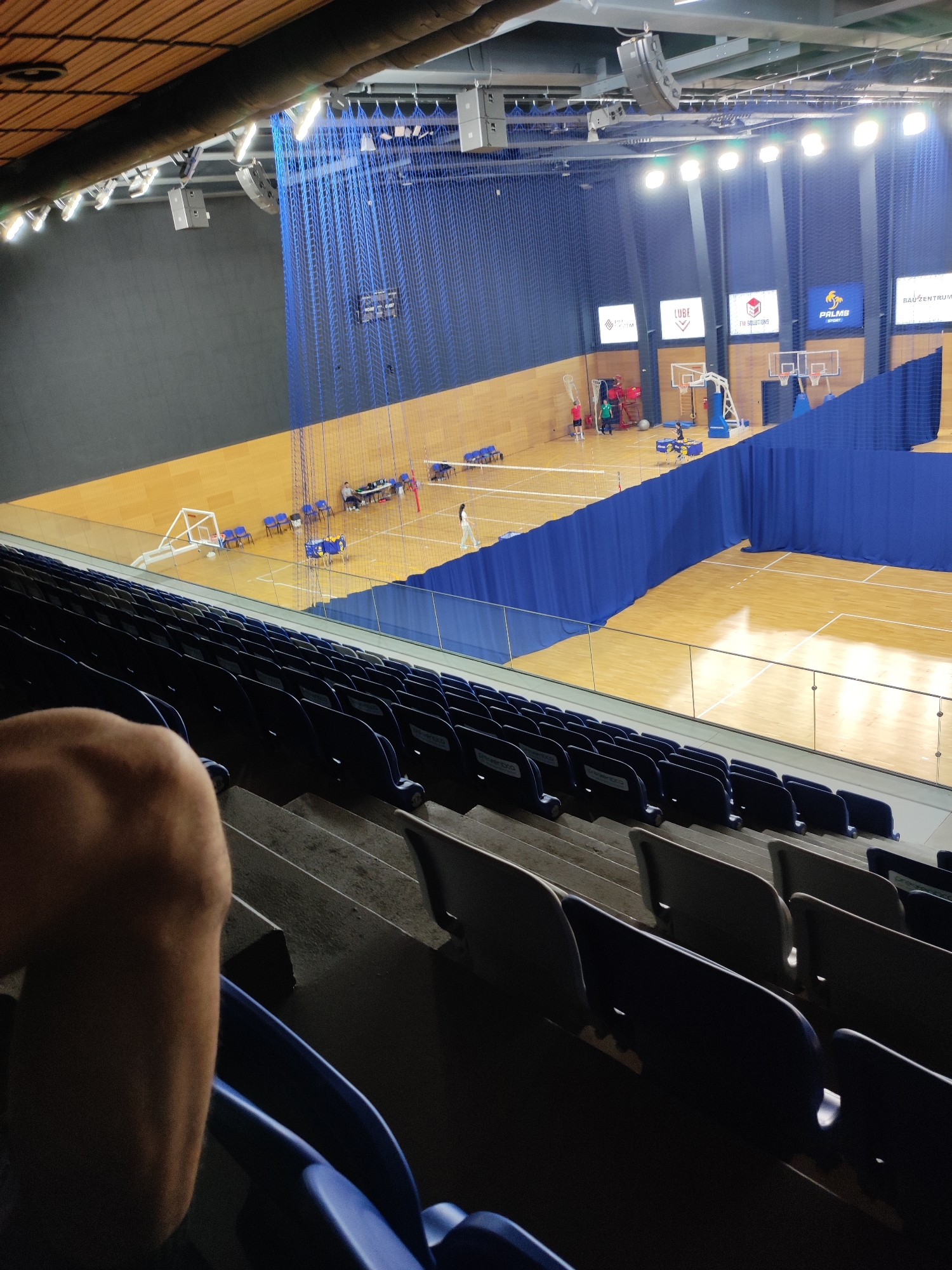 The image depicts the interior of a sports hall, likely a gymnasium, with basketball courts marked on the wooden floor. Several basketball hoops are visible, some with backboards. A blue net separates sections of the hall, and there are also blue curtains used as dividers. Spectator seating is arranged in tiered rows, with dark blue seats. The lighting suggests an indoor environment. In the background, several large screens or banners are mounted on the wall, some displaying logos and text. There are a few people visible on the court, some appearing to be players or coaches. The overall impression is of a modern sports facility. The provided context indicates this location is in Sofia, Bulgaria.