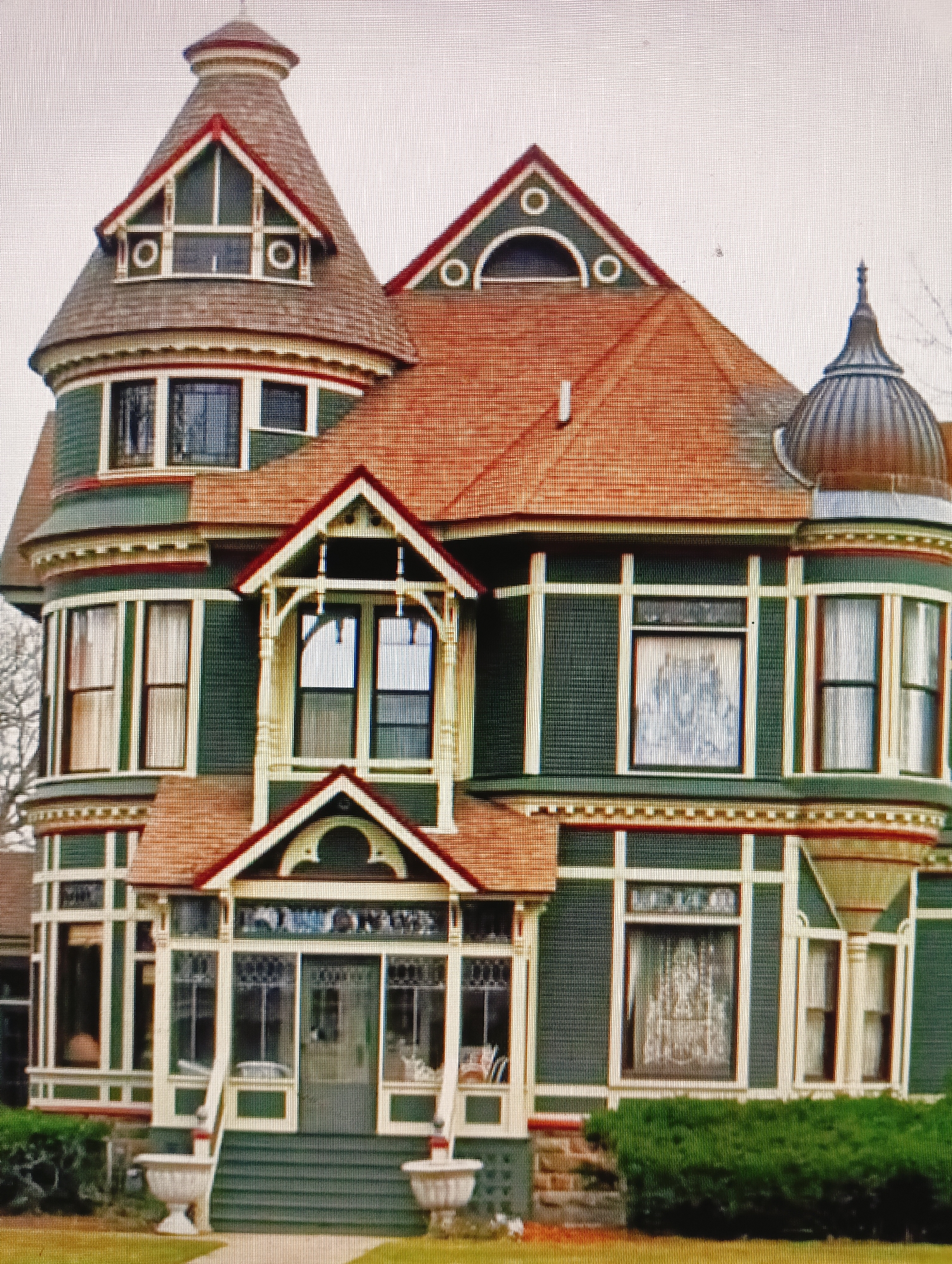 The image is a close-up, eye-level, exterior shot of a large, ornate, multi-story house with a complex architectural design. The house is painted a dark green with contrasting white trim and red accents. It features several rounded turrets, bay windows, a prominent conical roof, and a domed cupola. The roof is covered in reddish-brown shingles. The windows are multi-paned, and some appear to have decorative coverings or curtains. The house is surrounded by green landscaping, including manicured hedges and grass. In the foreground, there are two decorative urns on pedestals flanking a set of green stairs leading to an entrance. The sky is overcast and appears a pale gray. The image does not depict any people or specific landmarks that would indicate its location in Sluck, Belarus.