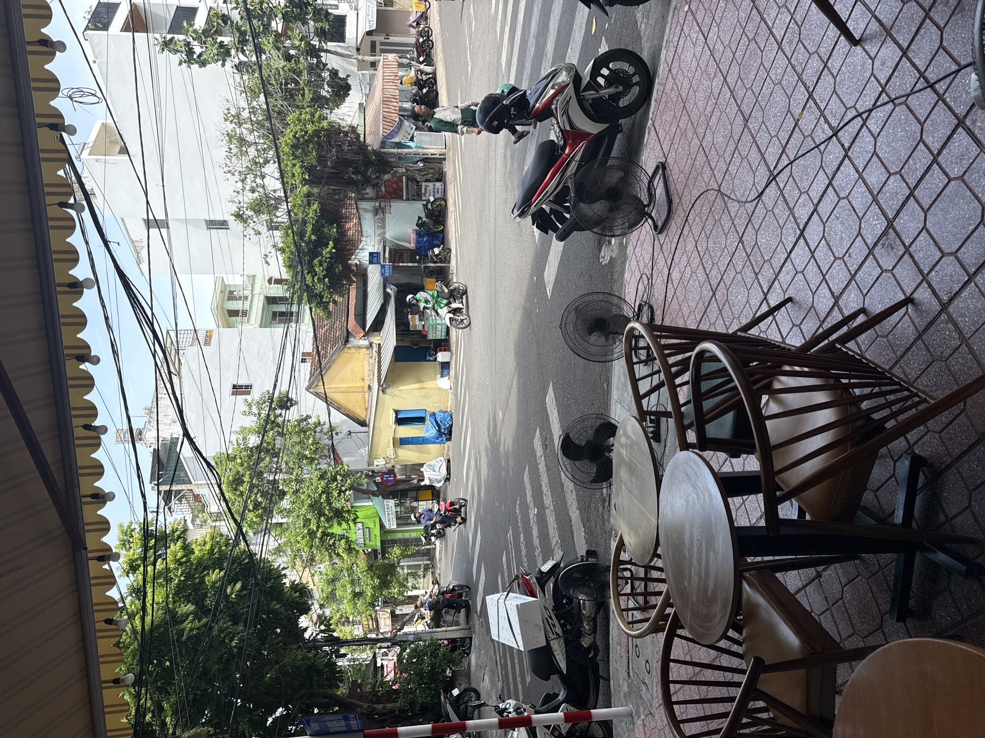 This is a street-level, eye-level shot of a street scene in Nha Trang Ward, Vietnam. In the foreground, two round wooden tables with four chairs each are positioned on a red tiled sidewalk. Three large, black oscillating fans are placed on the sidewalk, plugged in and facing the street. A motorcycle with two white boxes strapped to the back is parked on the sidewalk.
The street is paved and appears to be a multi-lane road with a crosswalk marked by white stripes. Several motorcycles are visible on the street, some in motion and some parked. People are seen riding motorcycles, with one individual wearing a green helmet and jacket. Pedestrians are also present, with one person in a green and white shirt walking across the crosswalk.
Buildings line the street, varying in architectural style and color. Some buildings are light-colored with balconies, while others are painted in yellow and have blue window frames. Lush green trees with dense foliage are interspersed along the street and between buildings, providing shade. Overhead, a canopy extends from a building, with a string of bare light bulbs hanging from it. A complex network of electrical wires is strung across the sky, a common sight in Vietnamese urban areas. The overall atmosphere suggests a busy, active street scene during daylight hours.