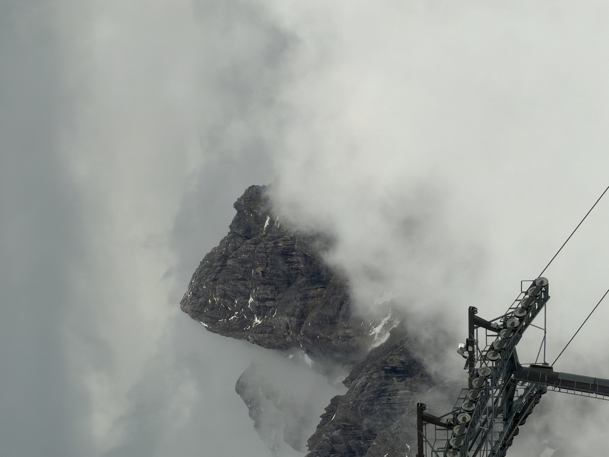 This is a vertical shot of a mountainous landscape near Garmisch-Partenkirchen, Germany, dominated by a thick cloud cover. In the upper-central portion, a prominent, dark grey, jagged rock peak emerges from the clouds, with sparse patches of white snow visible on its craggy surfaces. To the left and slightly below this main peak, other less defined mountain ridges are partially visible through the dense, white fog that envelops much of the scene. The sky above the mountains is uniformly grey and overcast. In the lower-left quadrant, a substantial metal structure, identified as a ski lift tower, extends diagonally upwards and to the right. This tower features multiple wheels or rollers designed to guide ski lift cables, one of which is visible stretching horizontally towards the right edge of the frame. The overall atmosphere is misty, moody, and devoid of direct sunlight. No people or animals are visible in the image.