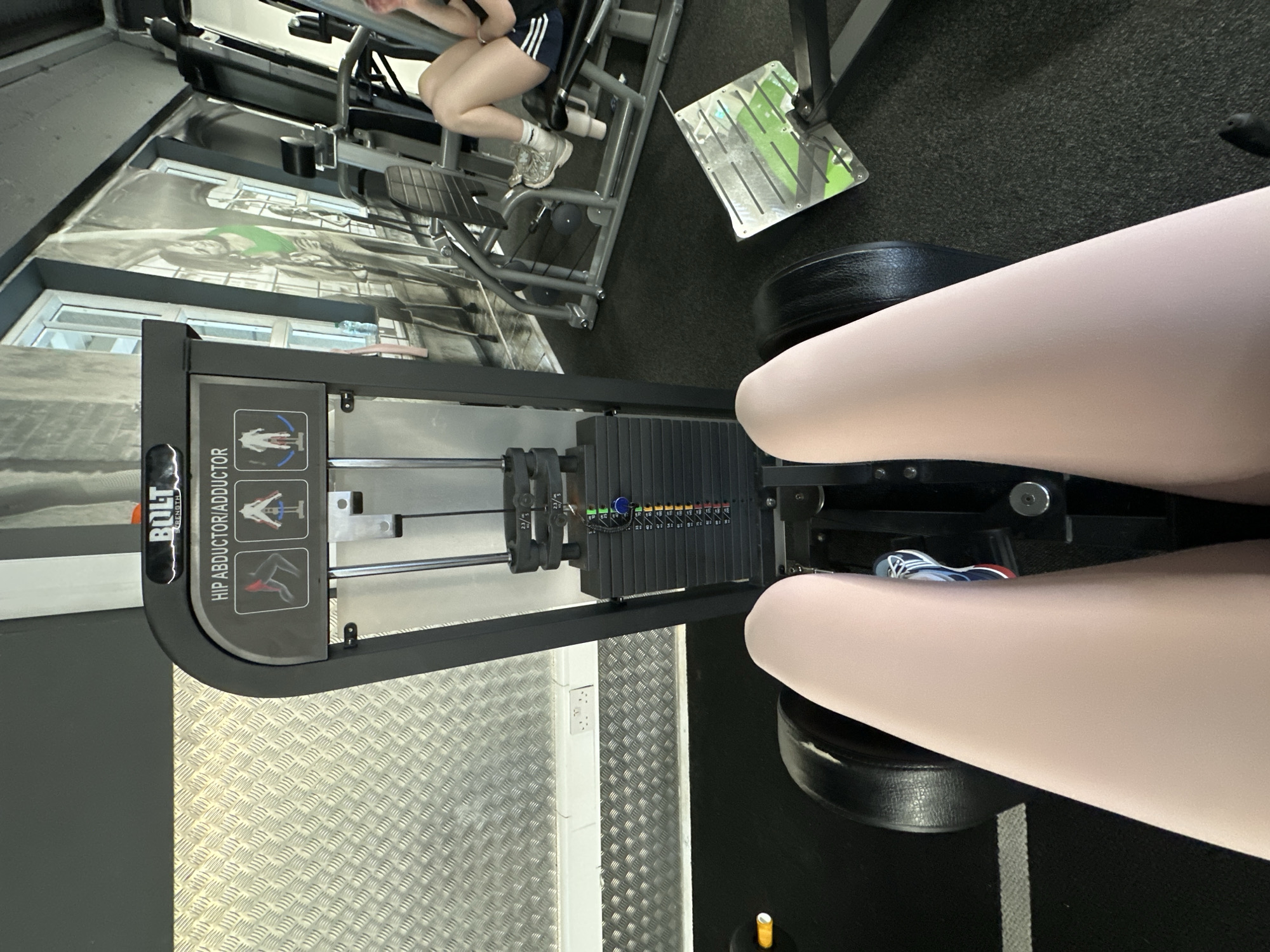 The image shows a first-person perspective of someone using a hip abductor/adductor machine in a gym. The machine is black and has a weight stack. The user's legs, wearing light-colored leggings and athletic shoes, are visible in the foreground. In the background, another person is visible using a leg press machine. The gym environment features gym equipment, a textured wall, and large windows. The gym is identified as being located in Carlow, Ireland.