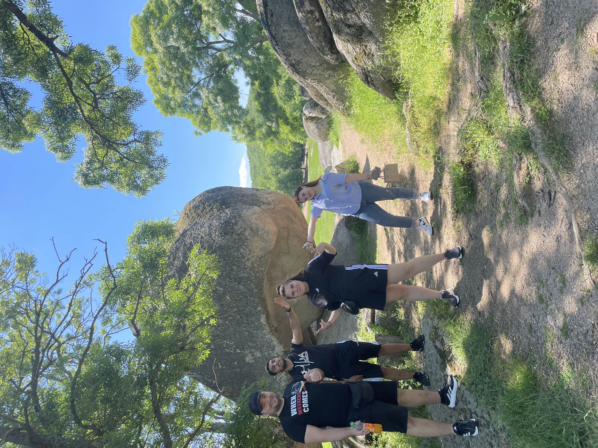 The image depicts a group of four people posing for a photo outdoors, likely in a park or natural area. The scene is bathed in sunlight, with a clear blue sky visible through the trees. The environment features large rock formations, lush green trees, and grassy areas along the ground.
In the foreground, there are three men dressed in dark-colored casual clothing, including t-shirts and shorts. The person in the front is holding a bottle of juice. The other one has a T-shirt with “WHEN WEEKEND COMES” on it. The third one has a T-shirt with “UNDER ARMOUR” on it.
A woman in a blue striped shirt and grey pants is standing next to the rock.
The background features a path leading into a more open area of the park, with green hills visible in the distance.