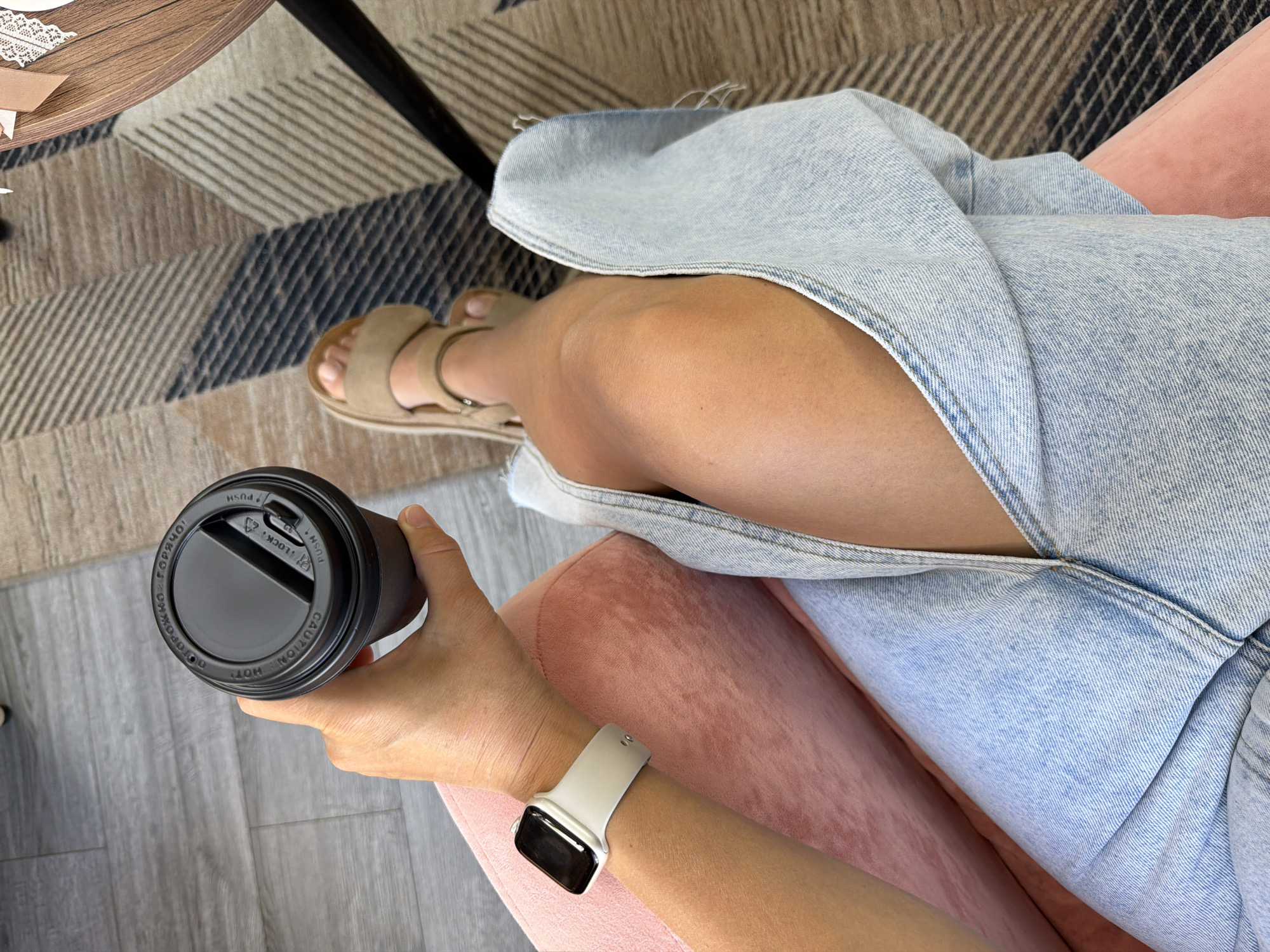 The media file shows a close-up of a person holding a black coffee cup while sitting on a pink chair. The person is wearing a light blue denim skirt and beige sandals. A white smartwatch is visible on their left wrist. The background features a carpet with a geometric pattern and wooden flooring. The scene appears to be indoors, likely in a cafe or a home. The lighting suggests it could be daytime. There are no specific cues to indicate the weather or a particular emotion. Text visible on the coffee cup lid includes "CAUTION HOT", "PUSH TO LOCK", and other unreadable text. The location context provided is Chaikovsky, Russia.