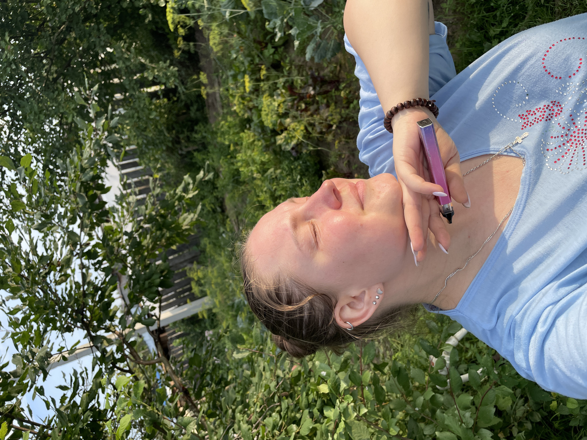A young woman is captured in a close-up, outdoor shot, bathed in the warm glow of what appears to be late afternoon sun. She is positioned in what seems to be a garden or yard, with lush green foliage and trees forming the backdrop. The environment suggests a rural or semi-rural setting, possibly in the vicinity of Балдейка, Russia, given the characteristic dense vegetation and hints of wooden fencing in the distance.
The woman has her eyes closed, her face turned slightly upwards and to the side, conveying a sense of peacefulness or enjoyment of the moment. Her hair is pulled back into a bun, and she is wearing a light blue t-shirt adorned with a patterned design of red and white embellishments. She is wearing a silver necklace with a small cross pendant and a bracelet made of dark wooden beads on her right wrist. Her left hand is raised, resting gently on her chin, and she is holding a pink and purple electronic cigarette device. The focus of the image is primarily on the woman's face and upper body, with the background slightly blurred, emphasizing her presence.