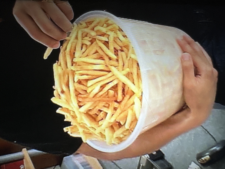 A pair of hands holds a large, slightly translucent bucket overflowing with golden French fries. One hand reaches into the bucket, pinching a single fry between its fingers. The bucket is tilted towards the viewer, showcasing the mass of fries within. The background is blurred, hinting at a setting with a black shirt and a glimpse of other objects on a table or counter. The image quality appears to be captured from a screen.