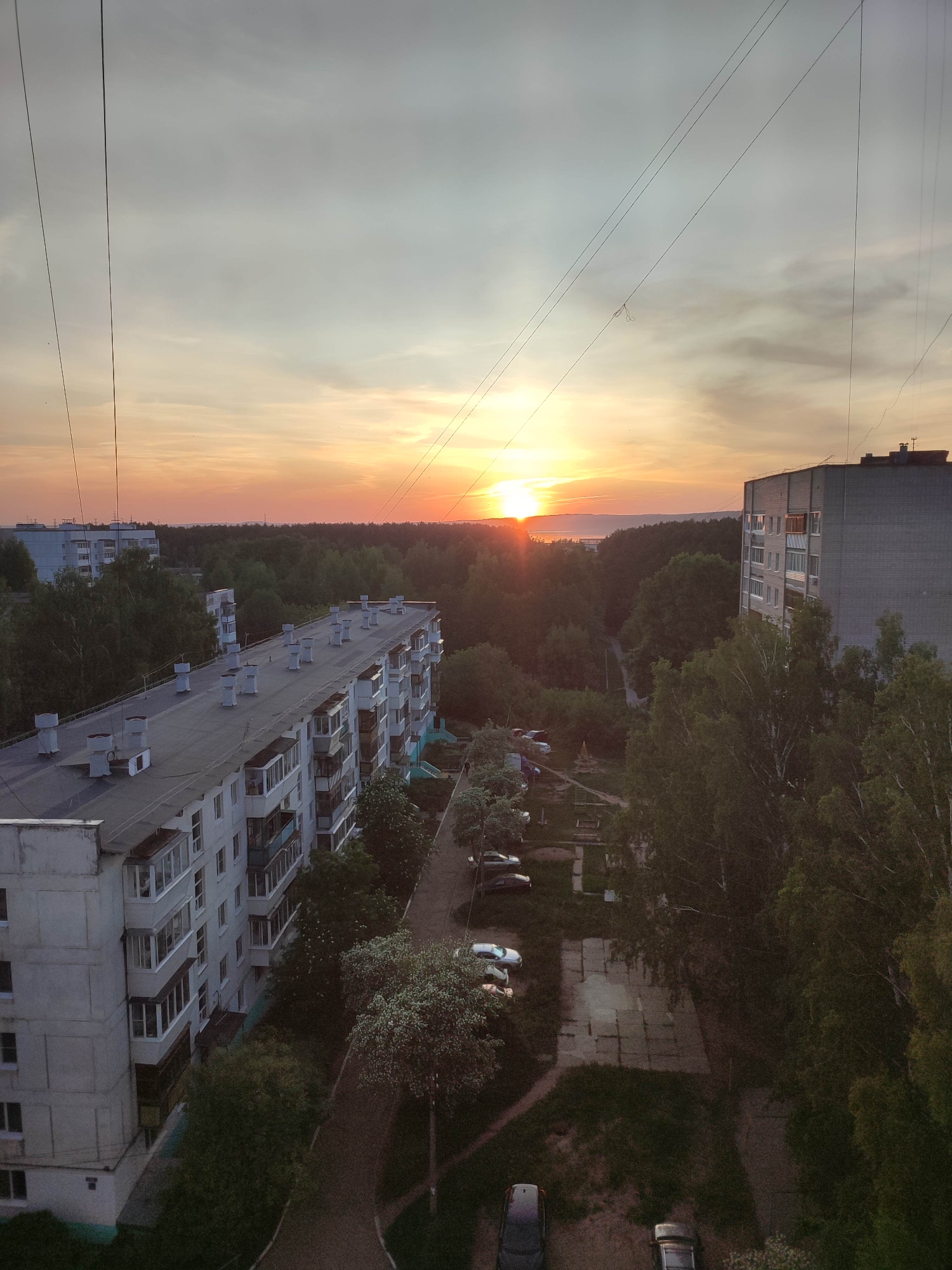 The image shows a sunset over a residential area in an urban location. The sky is a gradient of colors, with orange and yellow hues near the horizon where the sun is setting. The foreground is dominated by multi-story apartment buildings with flat roofs and rows of balconies. Trees are interspersed among the buildings and line the streets, suggesting a blend of urban and natural elements. A parking area with several parked cars is visible. In the distance, there is a line of trees and possibly a body of water on the horizon, situated beneath the setting sun. Power lines are strung across the top portion of the image. The scene appears calm and serene, captured during twilight.