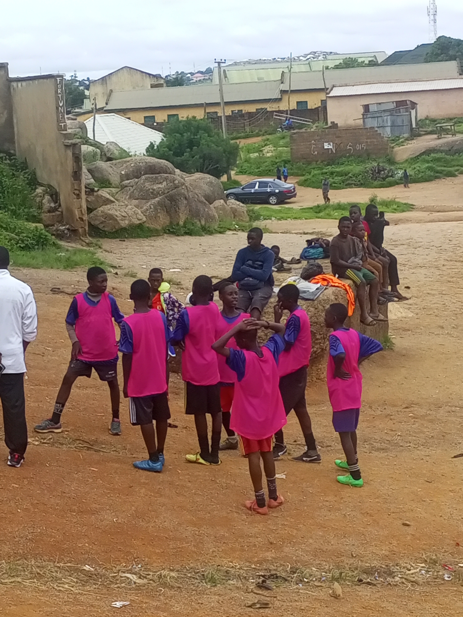 In Jos, Nigeria, a group of young boys, some wearing bright pink vests over blue shirts and shorts, are gathered on a dusty, uneven outdoor area. One boy on the left, in a white long-sleeved shirt and black pants, stands facing away from the camera. A few boys are seated on a concrete ledge in the background, some with their arms crossed, others looking towards the group. In the distance, several buildings with corrugated metal roofs are visible, along with a car parked on a paved road. Rocks and overgrown vegetation are scattered throughout the scene. The sky is overcast.