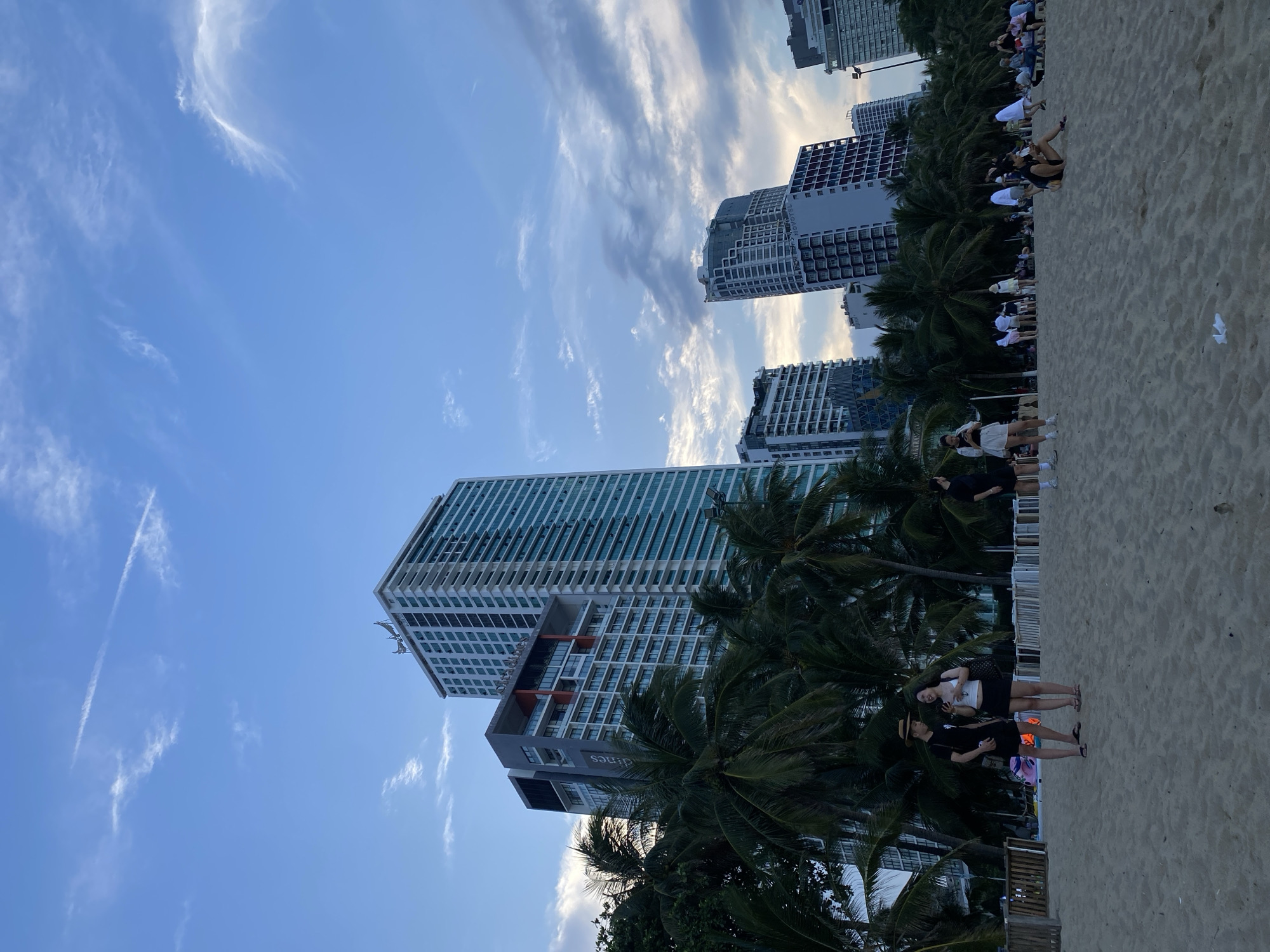 In Nha Trang Ward, Vietnam, a wide sandy beach is populated by numerous people and framed by lush palm trees and towering high-rise buildings. The sky above is a gradient of blue with wispy white clouds, suggesting it's either early morning or late afternoon.
In the foreground, the sandy beach is the primary surface, with footprints indicating recent activity. A few people are visible in casual attire, some standing and interacting, others sitting or walking. One couple in the mid-ground, a man in a black t-shirt, shorts, and a hat, and a woman in a white top and black skirt, are engaged in conversation. To their right, another man in athletic wear is standing, looking towards the water. Further to the right, a woman is sitting on the sand. In the background, a line of people is spread across the beach, some sitting in groups, others standing.
Behind the beach, a dense line of tall palm trees provides a tropical backdrop. Beyond the trees, the cityscape of Nha Trang is dominated by modern, multi-story buildings, including several distinct high-rise hotels and residential complexes. These buildings, with their numerous windows and varying architectural styles, create a stark contrast between the natural environment and urban development. The overall scene captures a lively beachside atmosphere with a prominent urban backdrop.