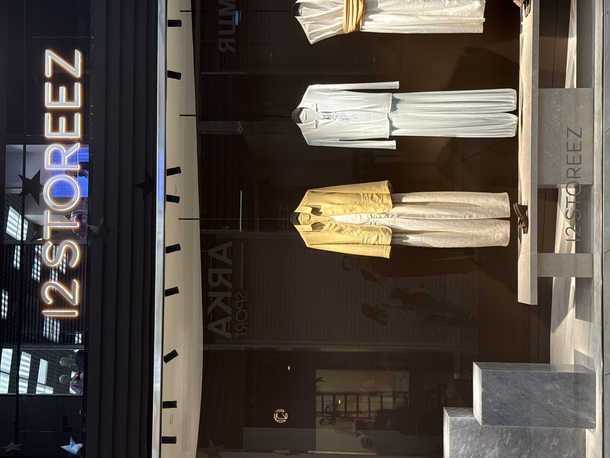 The image depicts a storefront with clothing displayed in the window. The text "12 STOREEZ" is prominently displayed vertically on the left. The name is also visible vertically on the right. The storefront is located in Perm, Russia, as "Perm Arka" is written backwards in the window. Three outfits are displayed in the window. The outfits consist of a white dress with a tan belt, a long-sleeved white shirt with a white skirt, and a gold jacket with a beige skirt. Three gray stone blocks are located on the bottom right corner of the image.