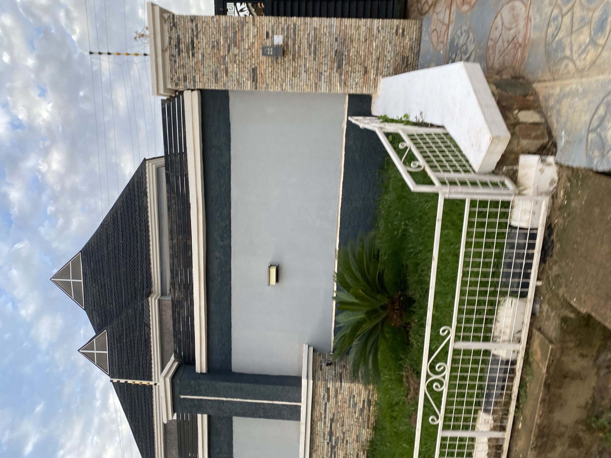 This image captures the exterior of a residential property in Bar Jirgi Summa, Nigeria. The focus is on a large, modern house with a dark, pitched roof featuring triangular dormers. A prominent grey wall, bordered by dark grey trim, extends across the scene, with stone-clad pillars at either end. A white metal fence with decorative scrollwork encloses a small patch of bright green grass with a sago palm. Electrical fencing is visible along the top of the wall and a brick pillar. The ground in the foreground is paved with patterned tiles and a concrete walkway. The sky above is partly cloudy.