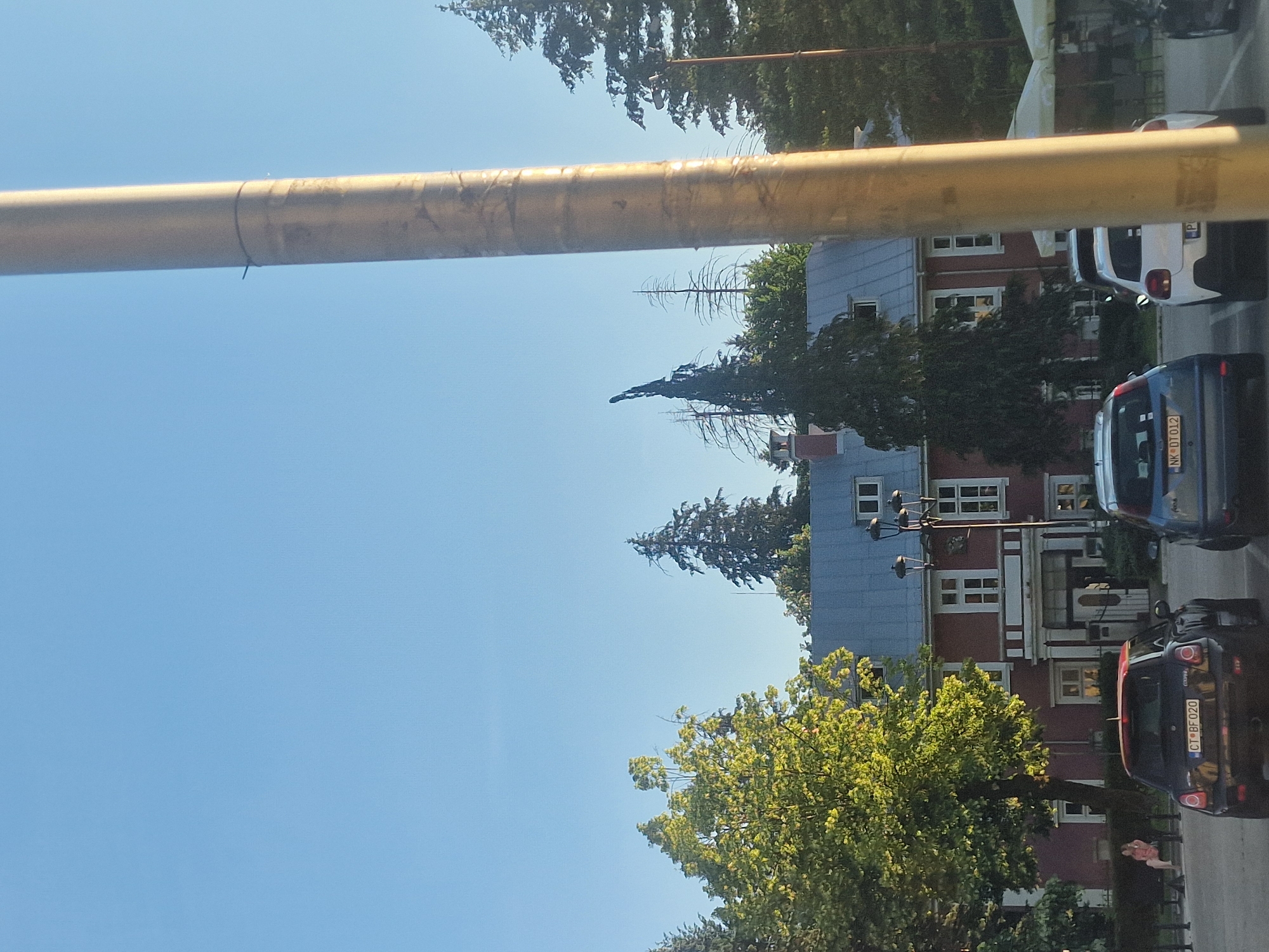 The image captures a street scene in Cetinje, Montenegro, on a clear day. In the foreground, a large, textured, light-colored pole dominates the right side of the frame, obscuring a portion of the background. In the mid-ground, a dark-colored Mini Cooper with license plate CT BF020 is parked in a lot, with a blue car and a white car to its right. Behind the cars, a long, red building with white-framed windows and a grey, sloped roof is visible. Several tall evergreen trees and deciduous trees with green foliage are scattered throughout the scene. A person is seated on a bench to the left of the red building. In the far background, the sky is a clear, light blue.