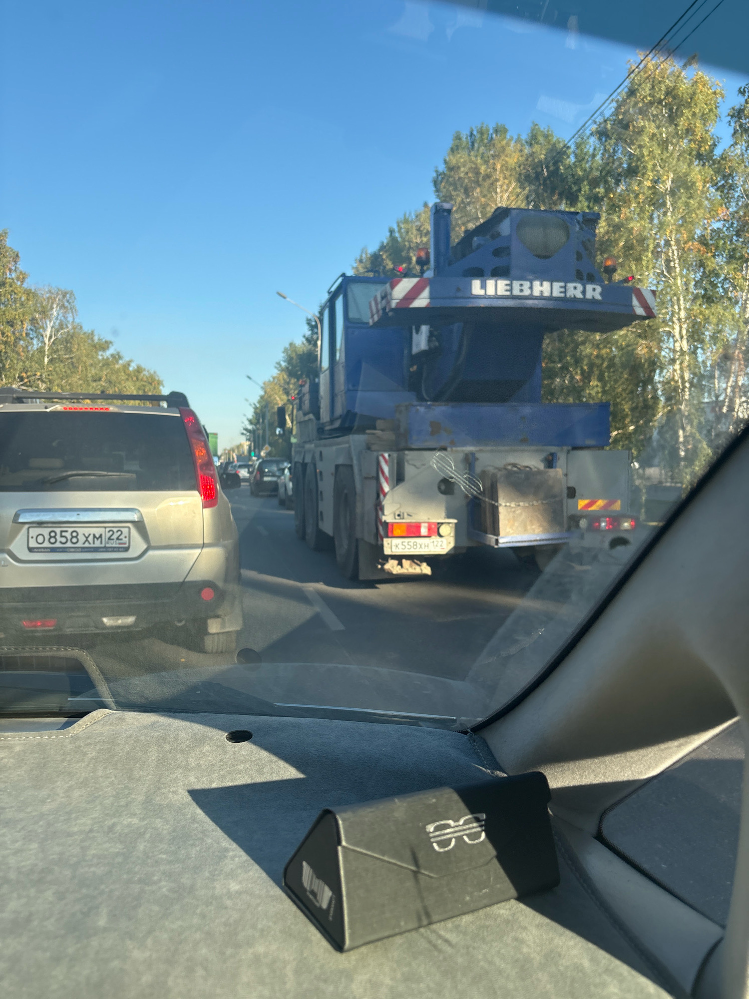 The image, taken from inside a vehicle, shows a traffic jam on a sunny day in Barnaul, Russia. A large blue Liebherr crane is the most prominent object, dominating the center of the frame. The crane is mounted on a truck chassis and appears to be in transit, as it is part of a queue of vehicles. To the left of the crane, the rear of a silver SUV is visible, displaying a Russian license plate that reads "O 858 XM 22". The SUV is also part of the traffic jam. In the background, trees line the road, suggesting an urban or suburban setting. The sky is clear and blue. The interior of the vehicle from which the photo is taken is partially visible, with the dashboard and a black case on it in the foreground.