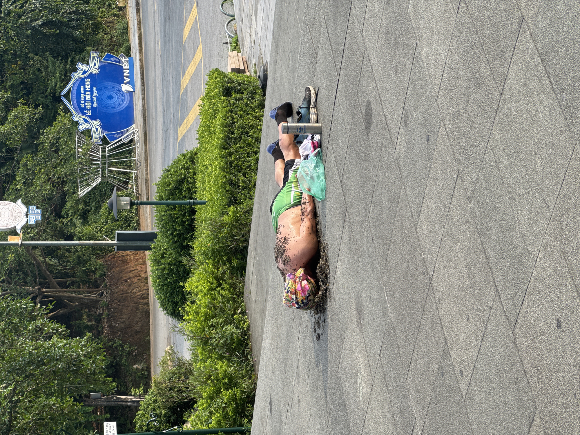 In the city of Hy Cương, Vietnam, a person is lying on their stomach on a paved walkway. The individual is wearing a green shirt and black shorts, with blue socks and black shoes. A colorful headscarf is on their head, and dirt and debris are scattered around them. Next to the person are a green plastic bag, some clothing items, a water bottle, and a shoe. In the background, there is lush green vegetation, a street with yellow markings, a lamppost, and a large blue sign with Vietnamese text and the VNPT logo. The sign appears to be promoting an event. The scene is illuminated by daylight, casting shadows on the ground.