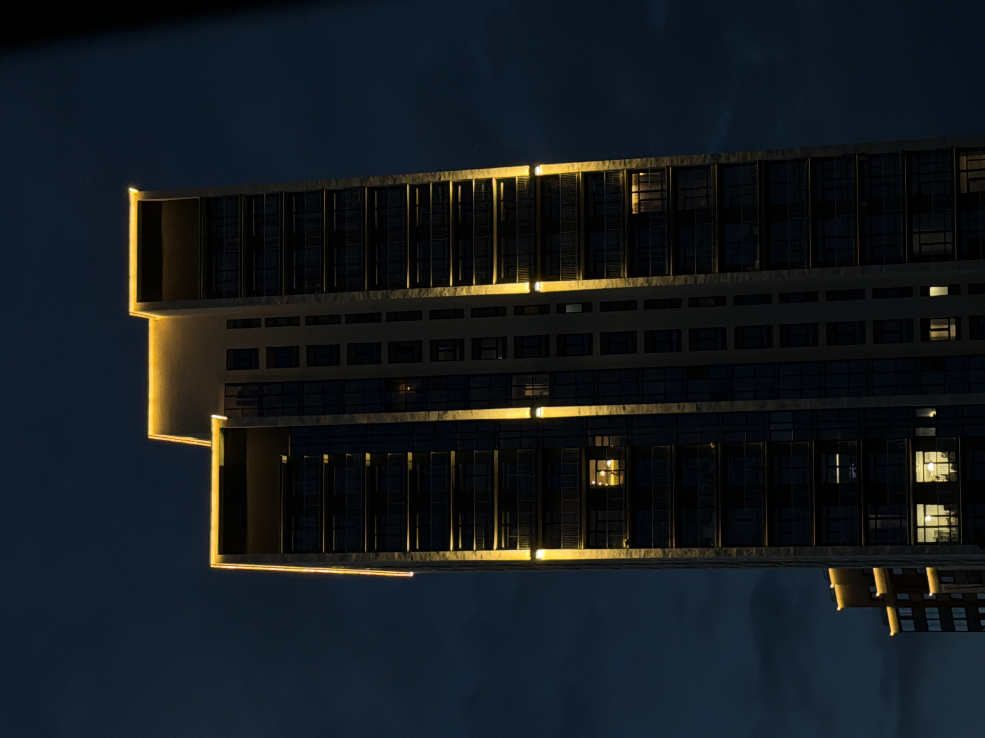 A tall, modern apartment building, illuminated by warm yellow lights along its edges and on several floors, stands against a dark, cloudy night sky. The building has a multi-story design with numerous balconies and windows, some of which show interior lights. To the left, another building of similar architectural style is partially visible, also featuring yellow exterior lighting. The image appears to be taken in the city of Yekaterinburg, Russia, likely in a contemporary urban district. There are no people or specific actions depicted in the scene.
