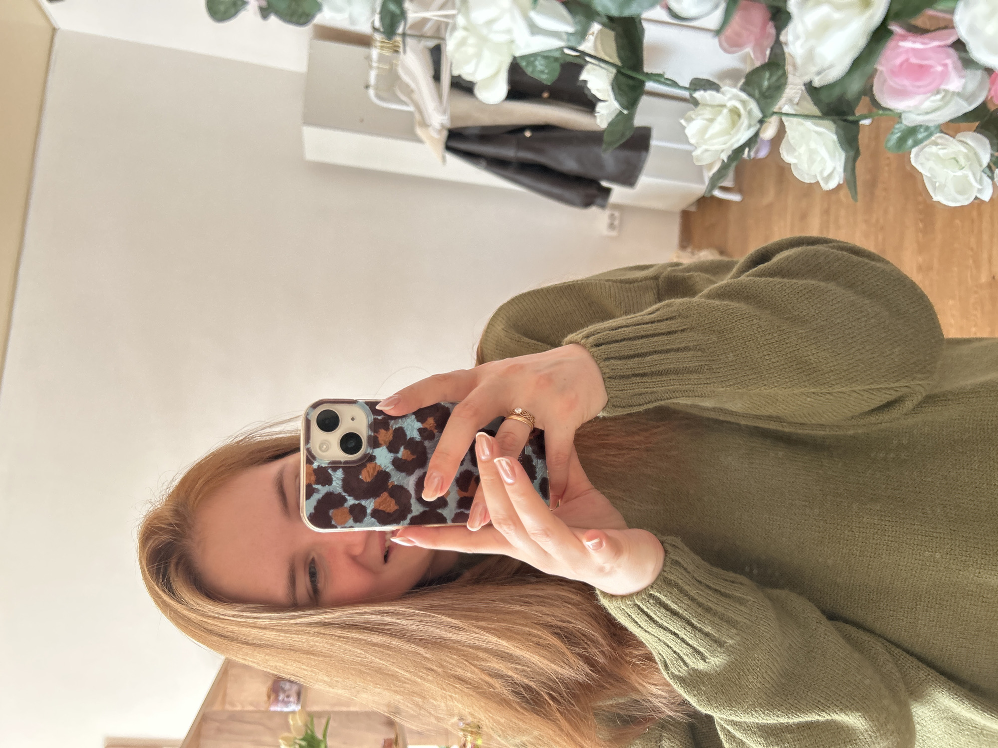A woman with long, light brown hair is holding a smartphone with a blue and brown leopard print case in front of her face, seemingly taking a selfie in a mirror. She's wearing a green sweater. In the background, there's a white wall, a clothing rack with a brown jacket hanging on it, and a decorative floral arrangement of white and pink roses. The location is not identifiable as the city of Chaikovsky, Russia, based on the visual information provided.