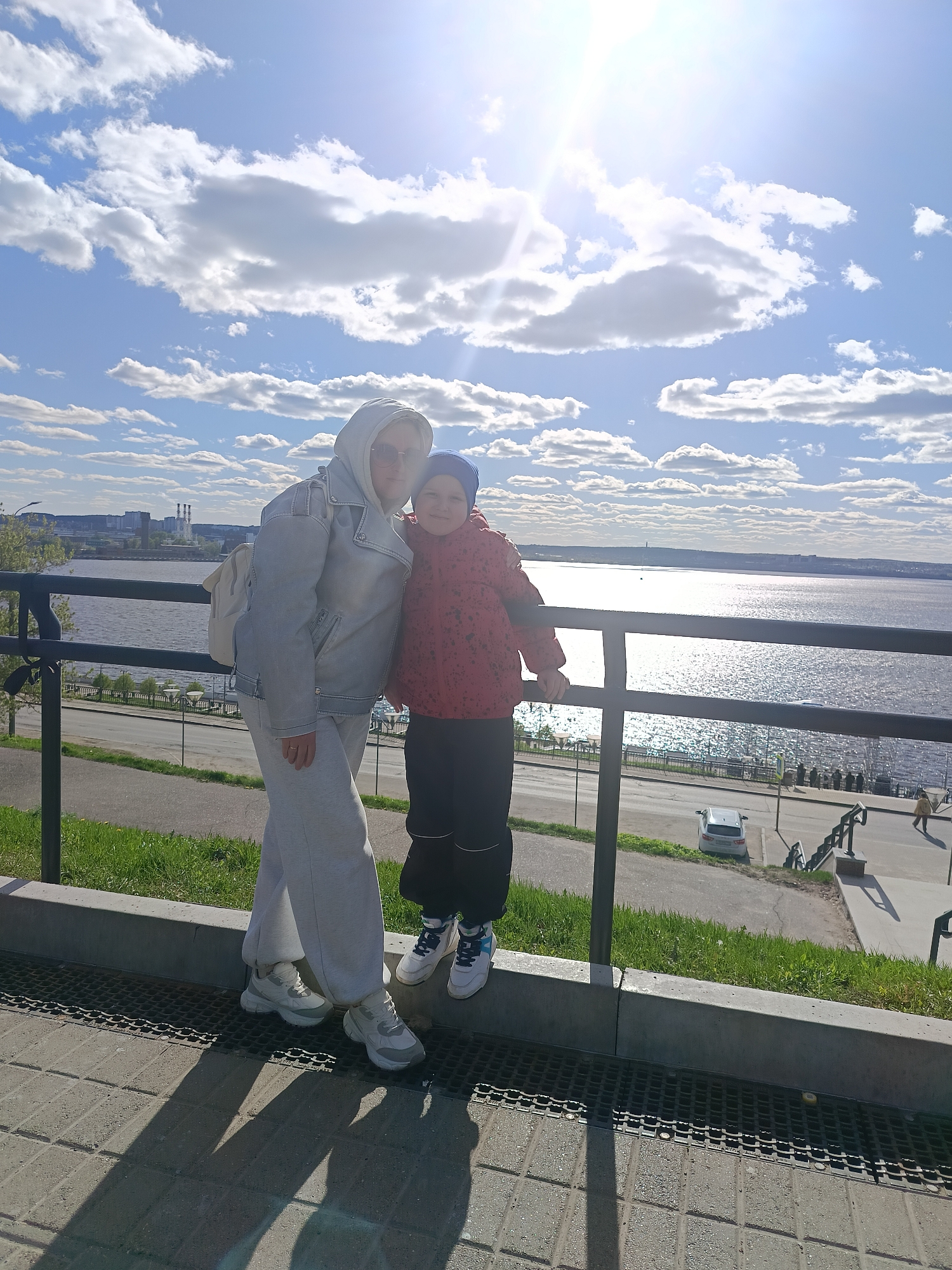 Two individuals, an adult and a child, are positioned beside a black metal railing, overlooking a body of water. The adult wears a light-colored hooded jacket with matching pants and white sneakers. A white backpack is slung over their shoulder. The child wears a red jacket, dark pants, white sneakers with blue accents, and a blue cap.
The backdrop consists of a wide river reflecting sunlight, a bright sky with puffy clouds, and the cityscape of Izhevsk, Russia. Visible elements of the cityscape include buildings and what appears to be a power plant with tall smokestacks.
Below the railing, a paved walkway, a grassy area, and a lower level with stairs and a parked white car are seen. People walk on the lower level. The ground where the individuals stand is paved with grey blocks. The scene is bathed in daylight, suggesting a sunny day.