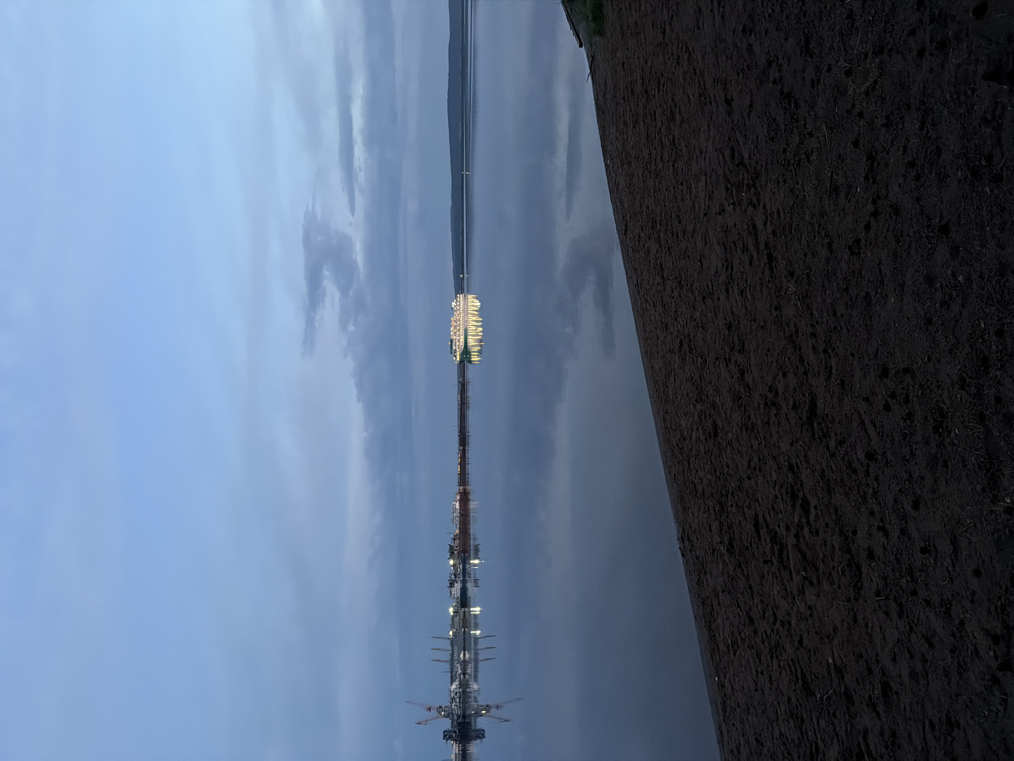 The image depicts a waterfront scene in Chaikovsky, Russia at dusk. A dark, textured embankment dominates the right side of the frame. A body of water stretches across the middle, reflecting the sky, which is overcast with a mixture of blue and grey clouds. A long, thin pier extends into the water, illuminated by numerous lights. At the far end of the pier, a brightly lit structure is visible. Closer to the viewer, there is machinery or equipment on the pier.