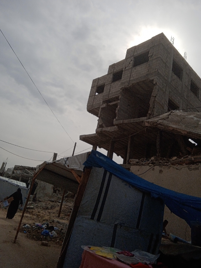 This image captures a scene of extensive destruction in Khan Yunis, Palestinian Territory, under an overcast sky with the sun partially visible behind structures. In the foreground, a makeshift shelter constructed from corrugated metal panels and a blue tarp leans to the right, with a red fabric and colorful items resting near its base. Adjacent to it, another crude shelter with a white tarp roof is supported by wooden posts. The ground throughout the scene is uneven, covered in rubble, dirt, and scattered debris, including discarded clothing.
To the left, a person clad in a black abaya walks from left to right, carrying a light-colored object under their arm, navigating the debris-strewn path. Another individual is partially visible near the base of the foreground shelter. In the midground and background, multiple structures exhibit severe damage. Dominating the right side of the frame is a multi-story concrete block building, appearing to be four to five stories tall. Significant sections of its exterior walls are missing, revealing the interior spaces and structural components. The lower floors show substantial collapse, with fragmented concrete slabs and exposed rebar, while the upper sections retain more structural integrity despite visible damage. Power lines stretch diagonally across the upper left portion of the sky.