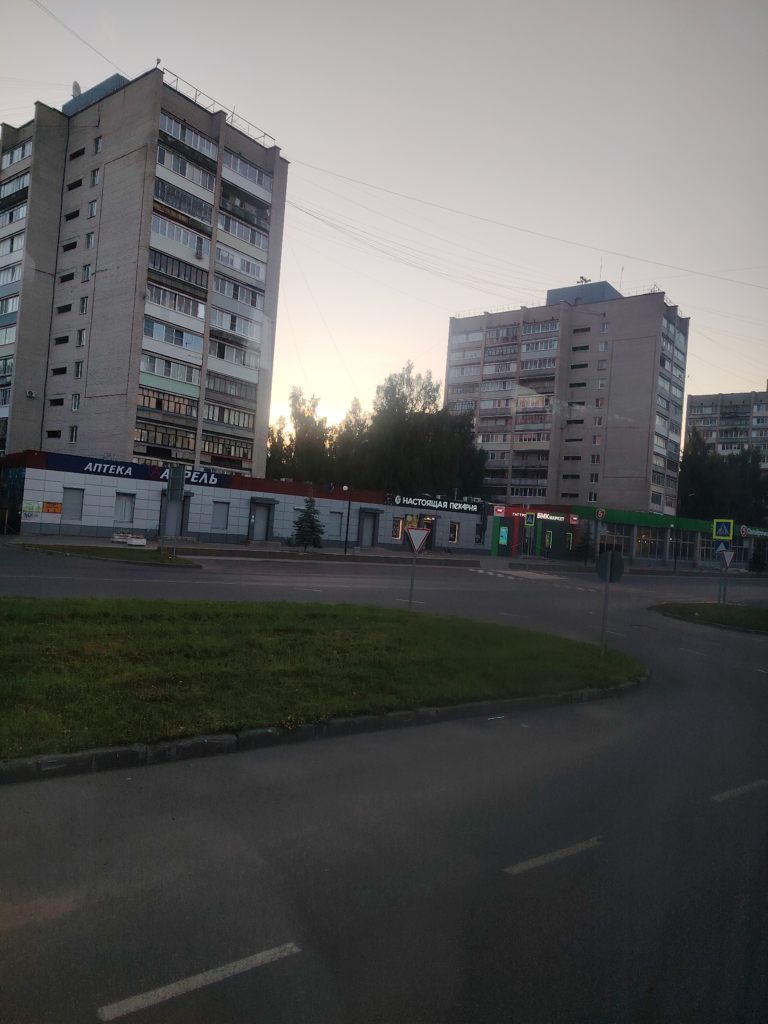 This is a daylight exterior, eye-level shot of a street in Konakovo, Russia. The scene features multi-story apartment buildings and commercial establishments lining a roadway. In the foreground, a paved road curves to the right, bordered by a strip of green grass and a concrete curb.
Dominating the left side of the frame is a tall, gray brick apartment building with numerous balconies and windows. Below it, a strip of commercial storefronts features signs for "АПТЕКА" (Pharmacy) and "А ПРЕЛЬ". Adjacent to this, further to the right, is another, slightly taller apartment building, also constructed of gray brick and featuring many windows and balconies. In front of this building are several shops with signs such as "НАСТОЯЩАЯ ПЕКАРНЯ" (Real Bakery), "БНК МАРКЕТ" (BNK Market), and others. A pedestrian crossing with yellow warning signs is visible on the right side of the road. The sky is overcast and pale. Several trees are visible behind the buildings, and power lines stretch across the sky. There are no people visible in the scene.