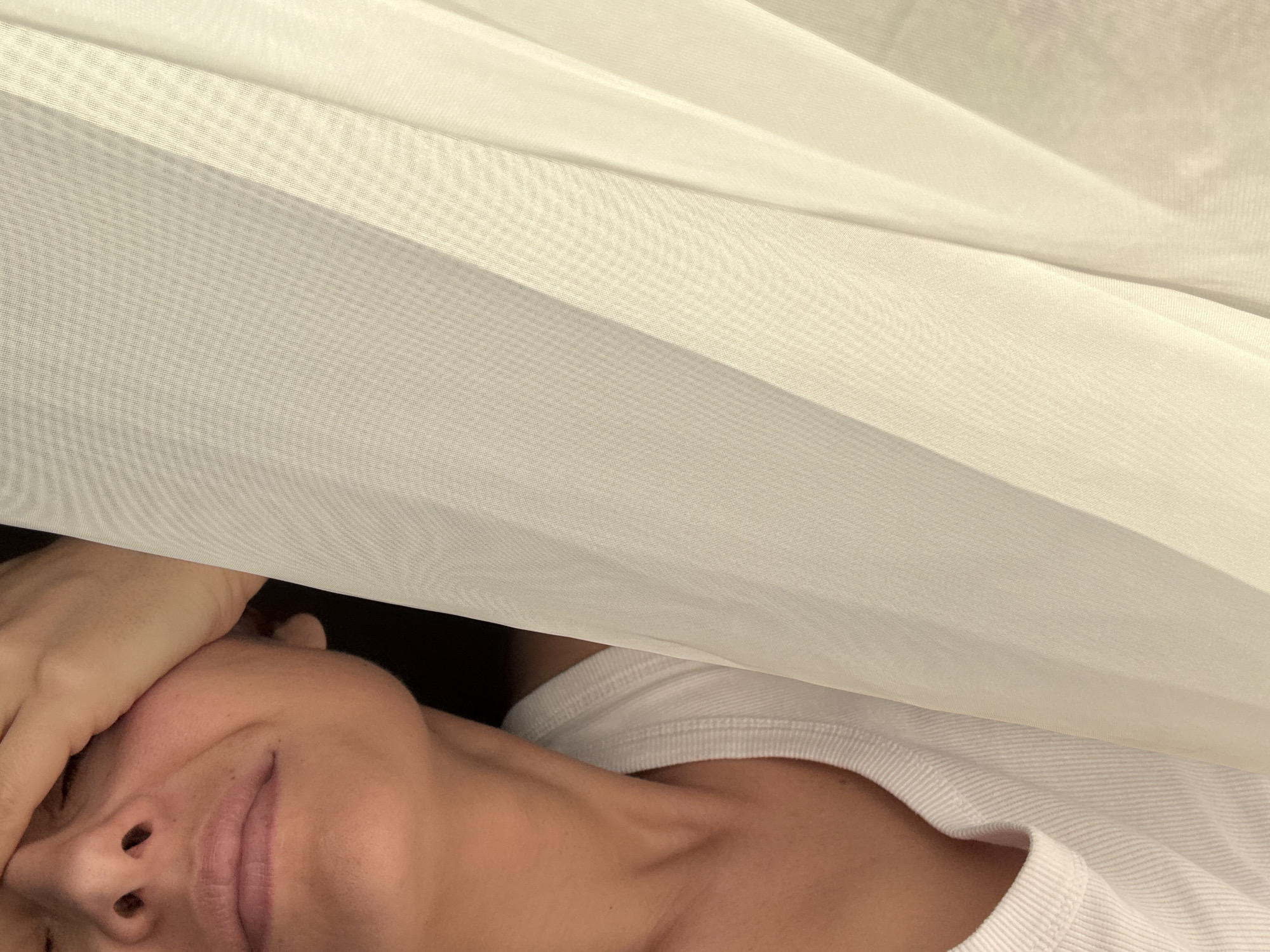 A close-up shot of a person lying down, possibly in a bed, partially covered by a thin, light-colored fabric or curtain. The person's face is visible, with their hand partially obscuring their forehead. They are wearing a white shirt. The background is dark and out of focus. The location cannot be determined from the image.