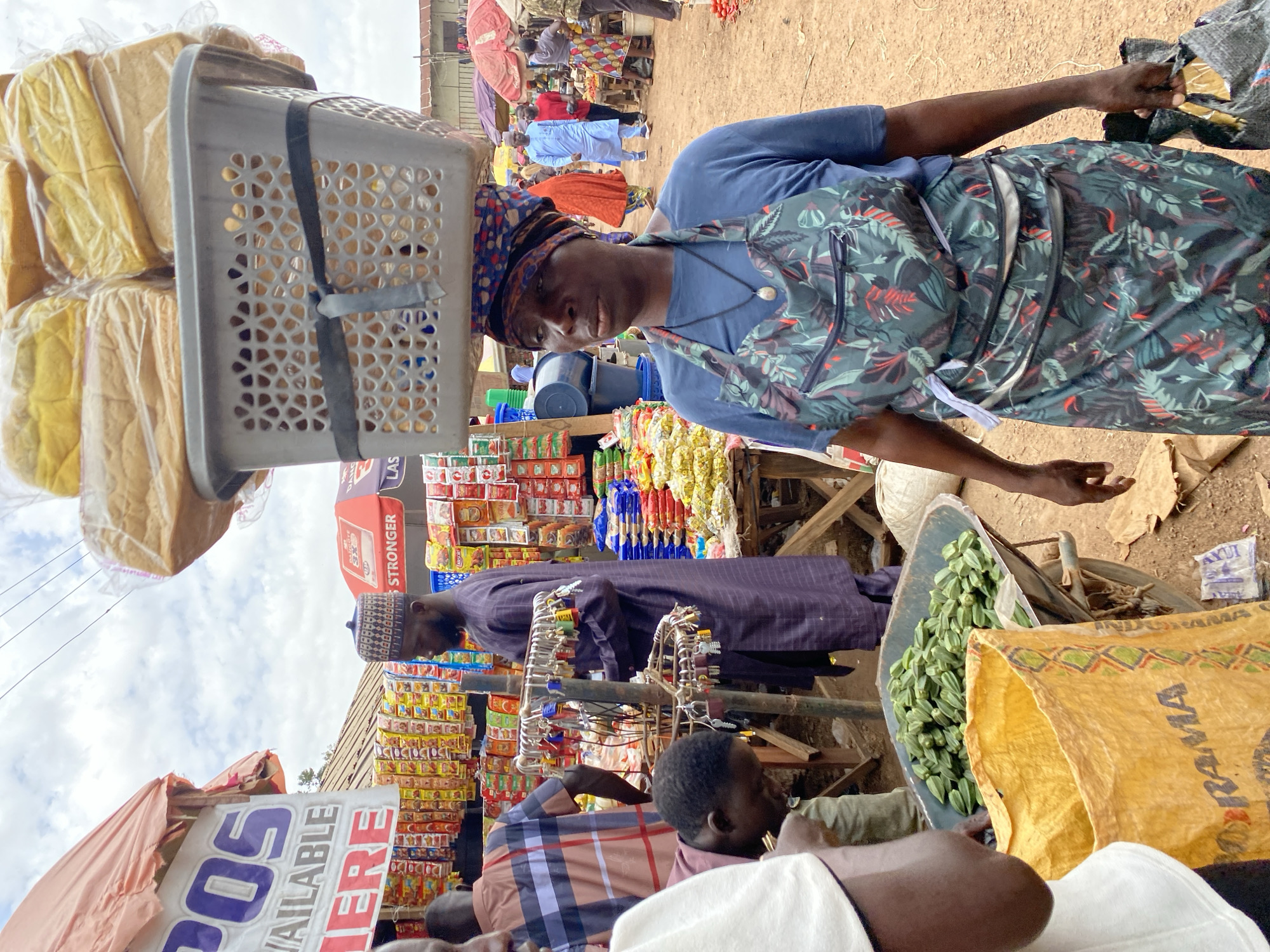 In Bukuru, Nigeria, a woman carrying a basket of bread on her head walks through a bustling marketplace. She wears a blue t-shirt and a floral apron, her head wrapped in a patterned scarf. In front of her, a wheelbarrow overflows with green okra, and a large yellow bag lies on the ground. Behind her, a man in a purple robe stands near a stall displaying various goods, including locks and chains. Other market-goers are visible in the background, some browsing stalls and others walking through the crowded space. The marketplace appears to be outdoors, with stalls set up under awnings and open to the sky.