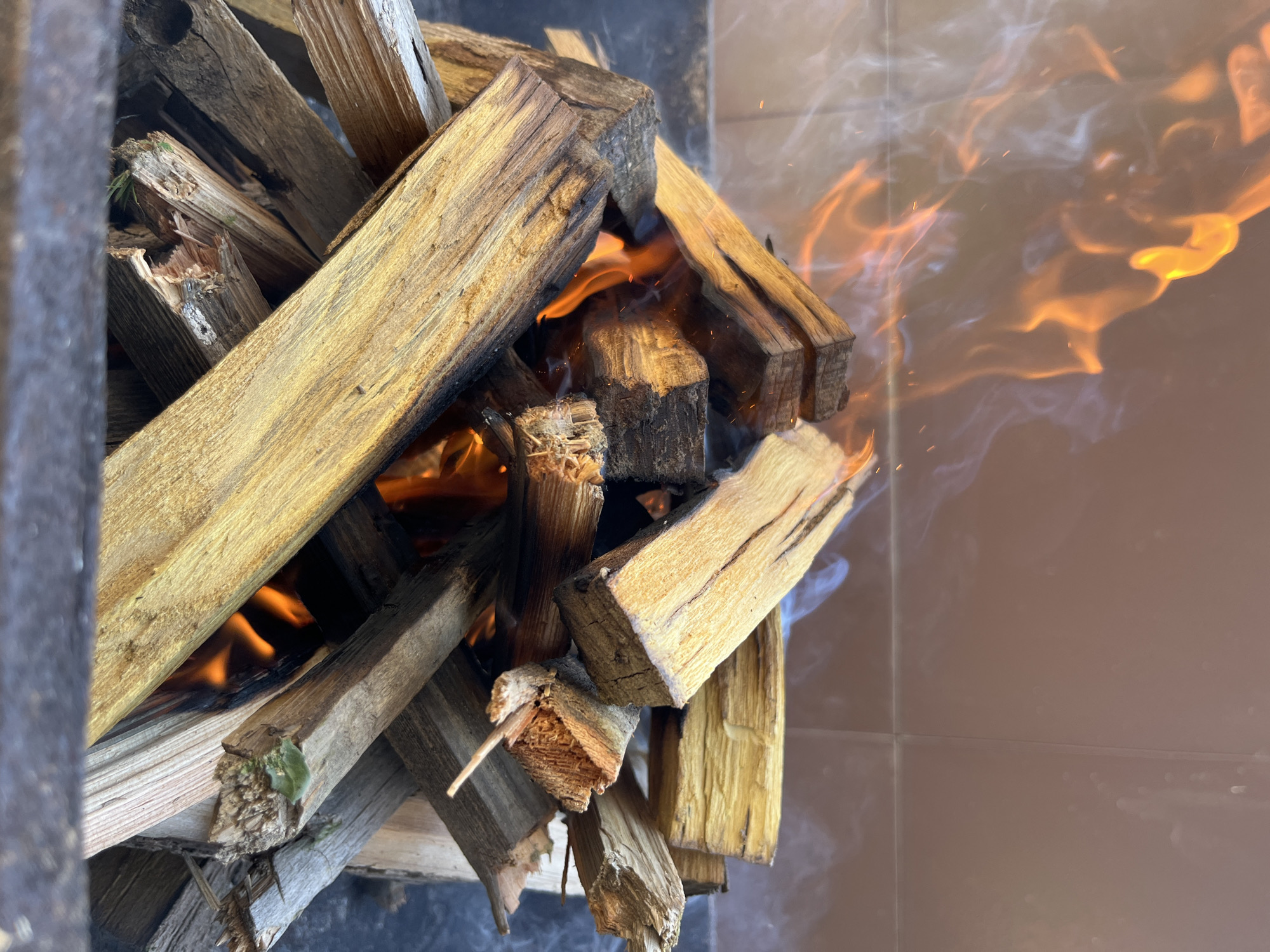 The image depicts a wood fire in a contained area. The fire consists of stacked wooden pieces that are actively burning with flames and smoke visible. The surrounding environment appears to be an indoor tiled area. The location is given as Kaspiysk, Russia but there is no confirmation of this in the image. There are no visible people in the shot.