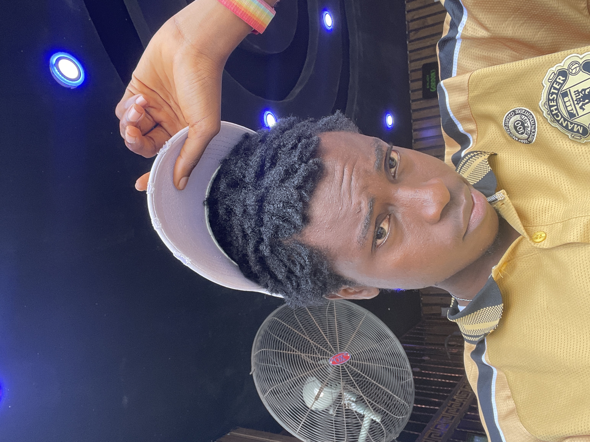 The media file features a close-up, rotated shot of a young Black man. He is holding a light-colored cap in his right hand, partially covering his dark, curly hair. He is wearing a gold collared shirt with black and white stripes along the collar and sleeves, and a Manchester United crest and a "100 years" patch are visible on the chest. A thin, multi-colored wristband is on his left wrist. His face is partially visible, with his eyes looking towards the camera. In the background, several blue LED lights are set into a dark, textured surface. Below the man, a large industrial fan with a metal grill is prominently displayed. The overall setting appears to be indoors or possibly a covered outdoor space at night, given the artificial lighting and the fan. There are no clear indications of weather conditions. Some text is visible on a tag on the shirt, including "GORDON". The mood conveyed is casual and relaxed. The location context is Nkpor, Nigeria.