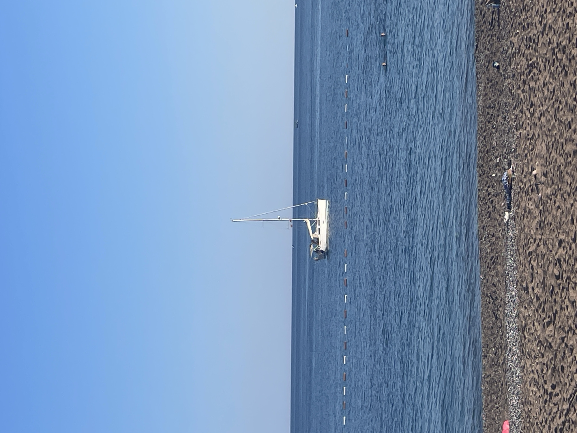 The image depicts a scene on a beach in Boreti, Montenegro. A white sailboat is anchored in the water, parallel to a line of white and brown buoys marking a swimming area. The water is blue and slightly choppy. To the right is the beach, characterized by dark sand and pebbles. The sky above is clear and blue.