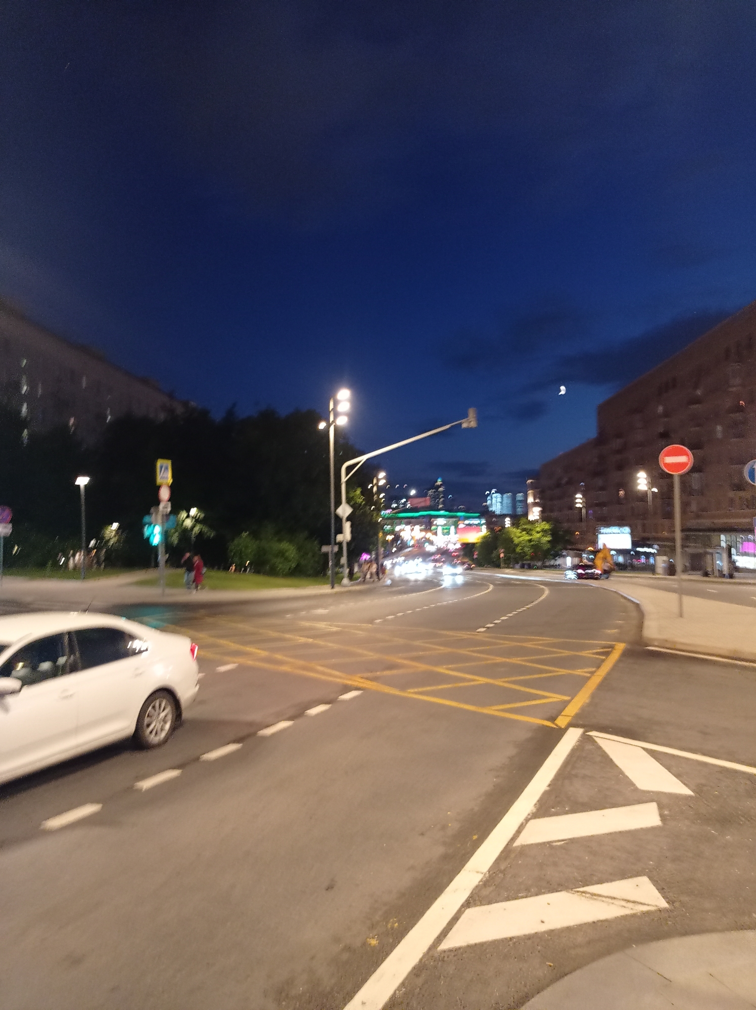 This is a photograph taken at dusk on a city street in Moscow, Russia. The image captures a road intersection with clear lane markings and pedestrian crossings, including a yellow box junction. A white car is visible on the left side of the frame, partially obscuring the road. Streetlights illuminate the scene, casting a glow on the asphalt and surrounding buildings. In the background, the silhouette of trees can be seen, along with several multi-story buildings, some with illuminated windows. Traffic lights and road signs are present, indicating a busy urban environment. The sky above is a deep blue, with a sliver of the moon visible. The overall impression is one of a typical evening scene in a Moscow neighborhood.