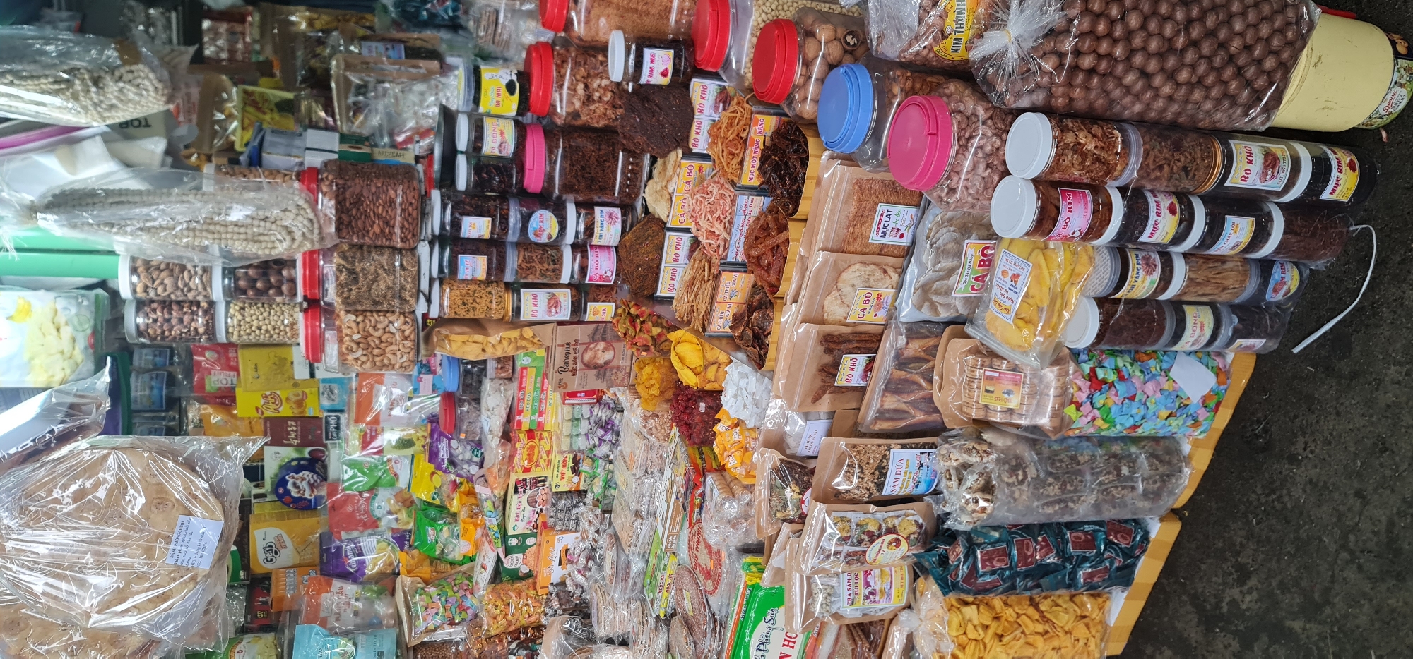 The image depicts a high-angle view of a market stall in Đà Nẵng, Vietnam, filled with a variety of dried goods and snacks. The items are arranged densely, and the main visible products are nuts, dried fruits, candies, and various types of dried meat. The packaging includes plastic containers, clear plastic bags, and cardboard boxes, many with labels in Vietnamese. The display suggests a diverse assortment of local specialties catering to tourists or local customers. The surface beneath the display has a mottled grey appearance.