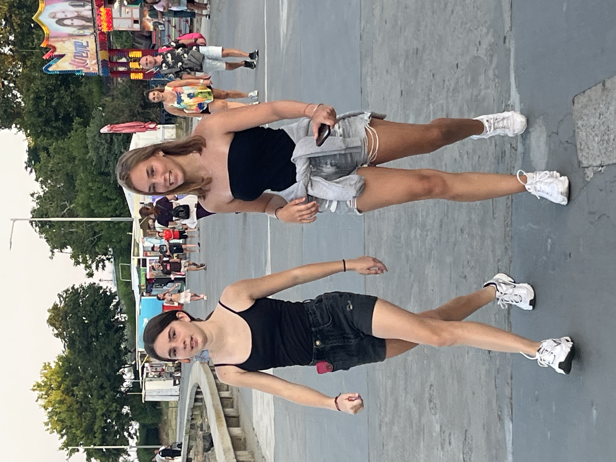 The image shows two young women standing outdoors, with a crowd of people in the background at what appears to be an amusement park or fairground. The woman in the foreground on the right is wearing a black tank top and dark shorts, with white sneakers. She has dark hair and is looking towards the camera with a neutral expression. The woman on the left is wearing a black strapless top, denim shorts, and white sneakers. She has blonde hair and is smiling and looking to the left. Her hair is tied around her waist with a grey hoodie. Behind them, there are several amusement rides, including one with "X-TREME" written on it. People are walking around, and the overall atmosphere suggests a lively event. The lighting indicates it is daytime, likely late afternoon or early evening, with soft shadows. The presence of trees and the overall setup suggest an outdoor setting in a recreational area. There are no strong indicators of weather, but it appears to be a clear day. The text "X-TREME" is visible on one of the rides. The scene appears to be set in Nesebar, Bulgaria.