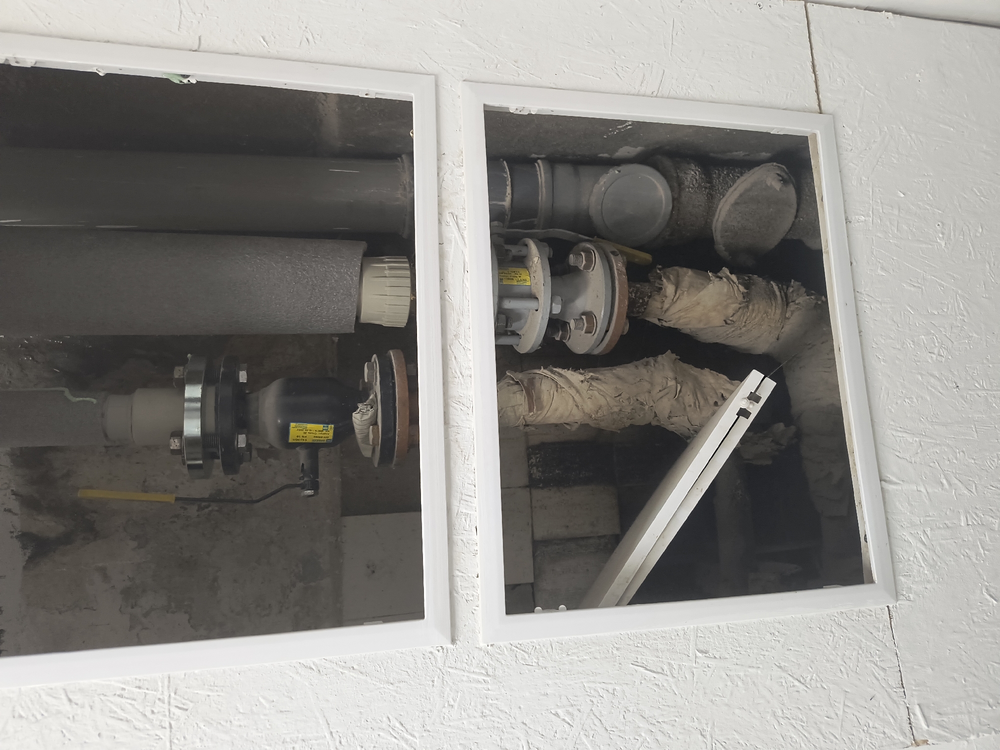 This is an exterior shot of a building's utility access panel in Rivne, Ukraine. The panel is divided into two rectangular sections. The top section shows plumbing with a dark valve and gray insulation. The bottom section reveals more plumbing, some of which is wrapped in a tan, fibrous material. A white panel is partially open in the bottom section, obscuring some of the view. The surrounding wall is textured and painted white.