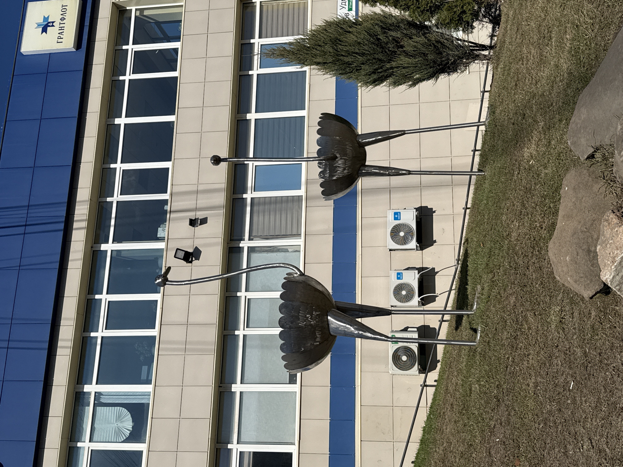 The image captures a view of the Izhevsk branch office of the Granitflot company in Russia. The building, featuring a combination of blue and beige tiled panels and multiple windows, is the backdrop for two metal sculptures resembling abstract birds or flowers. The sculptures are placed on a patch of grass next to a paved area where three air conditioning units are mounted on the building's facade. The sky is clear blue.