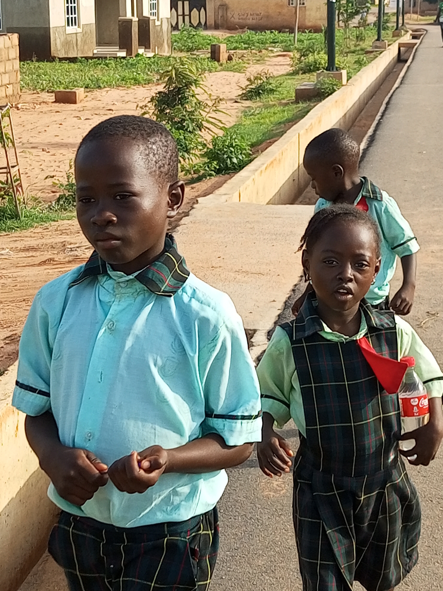 Three young children in school uniforms walk along a paved path in what appears to be a school environment in Tempure, Nigeria. The child in the foreground on the left wears a light blue collared shirt and dark plaid pants. Their hands are clasped in front of them. To the right, a younger child, also in a dark plaid jumper over a light green shirt with a red tie, holds a bottle of Coca-Cola. In the background, another child in a similar light blue uniform is partially visible. The environment features a sandy area with sparse green vegetation and some buildings with light-colored walls and dark windows. A low, yellow-colored wall runs along the side of the path.