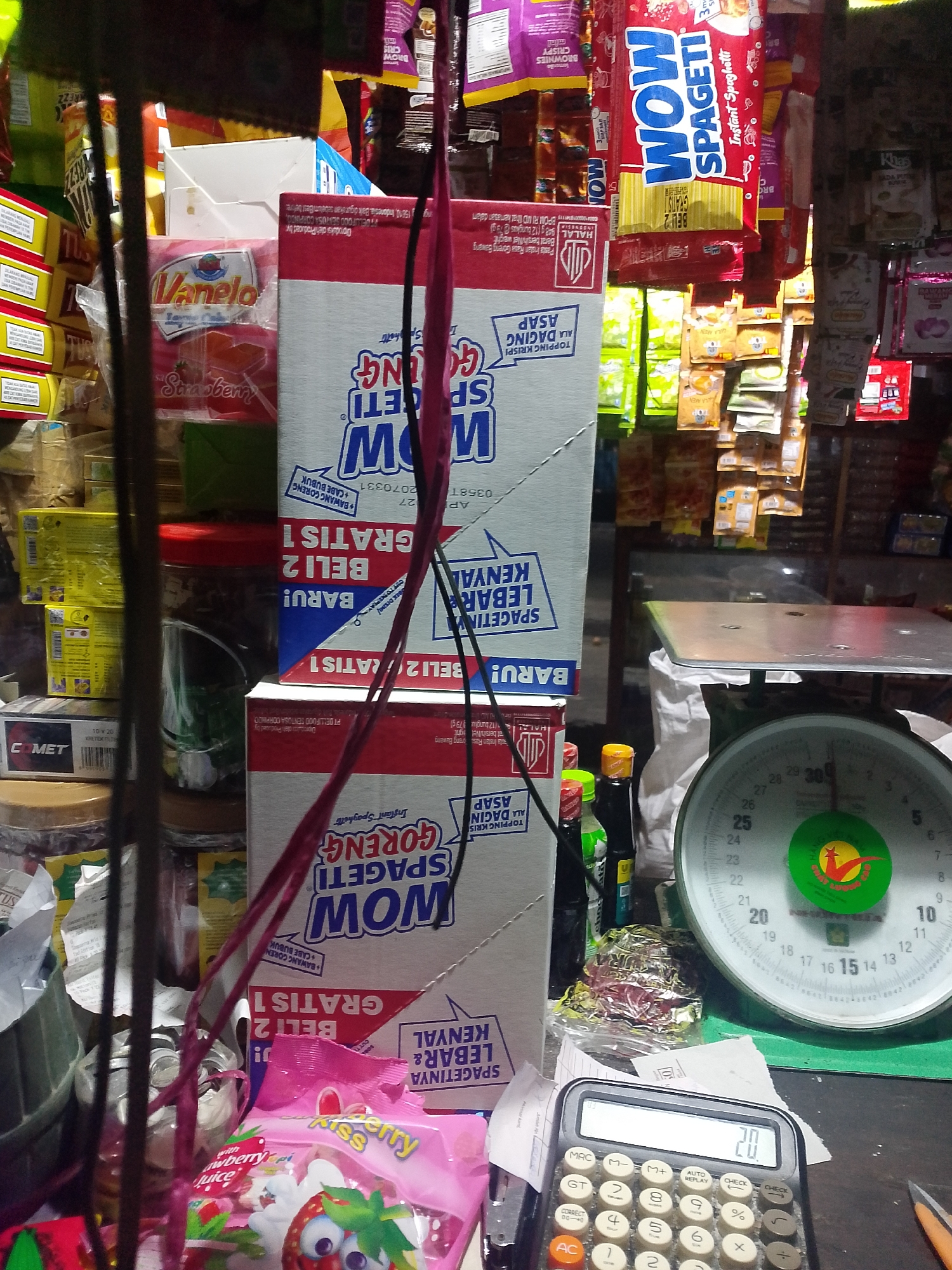 The image depicts a cluttered store shelf in Palembang, Indonesia, filled with various food products and merchandise. Prominently displayed are several boxes of "WOW Spaghetti" and "Vanelo Strawberry" snacks. Other items include bags of instant noodles, snacks, and what appears to be a traditional scale. The lighting is dim, suggesting an indoor environment, possibly a small neighborhood store or "warung." A calculator is visible in the foreground, suggesting recent sales activity. The overall impression is of a busy, well-stocked shop in a tropical climate.