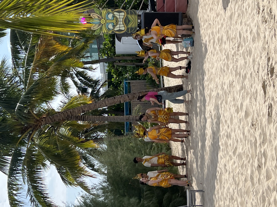 A beach scene features a row of dancers in yellow costumes with ornate headdresses. They stand on white sand next to palm trees, with a tiki-style decoration in the background. A person in pink stands observing. The dancers appear to be part of a performance, with the scene suggesting a tropical setting. The image captures the vibrant colors of the costumes and the sunny atmosphere of the beach.