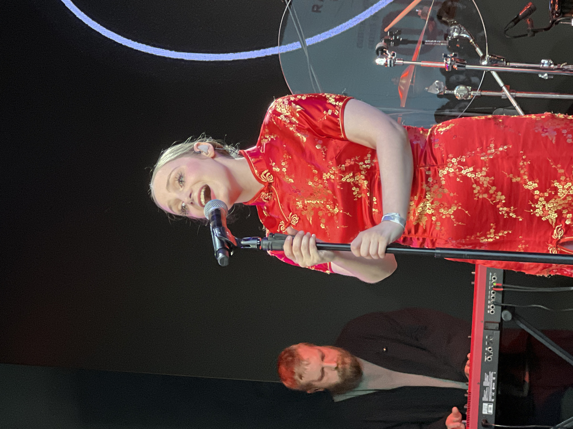 A woman in a red and gold floral print dress is holding a microphone and singing on a stage. She is fair-skinned with blonde hair. A man with a beard is visible below her, near a red keyboard. There are chrome drum stands in the background. The venue appears to be indoors, possibly a concert hall or club. The location is purportedly Saint Petersburg, Russia.