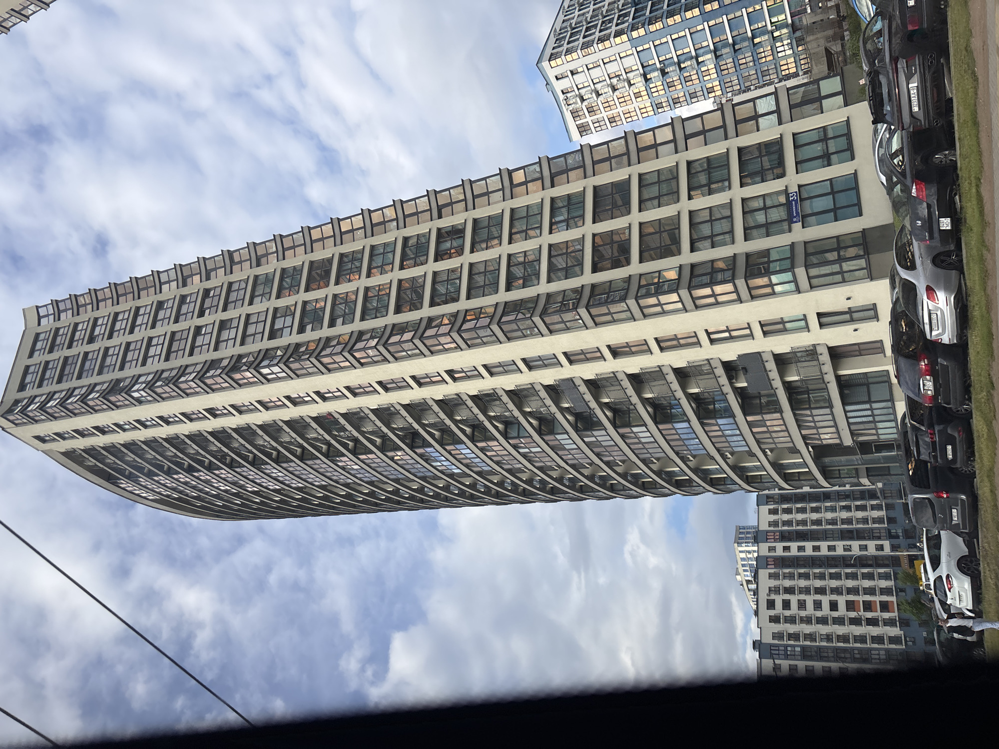 A tall, curved residential building with numerous windows and balconies dominates the frame, leaning to the right and upwards against a cloudy sky. In the background, to the right and further back, is another modern residential building with a blue facade and repeating window patterns. In the foreground, a line of parked cars, including a white hatchback, a silver sedan, and several dark SUVs and sedans, are visible. A person in a light-colored shirt and dark pants walks on the left side of the frame, near the parked cars. The environment appears to be an urban residential area.