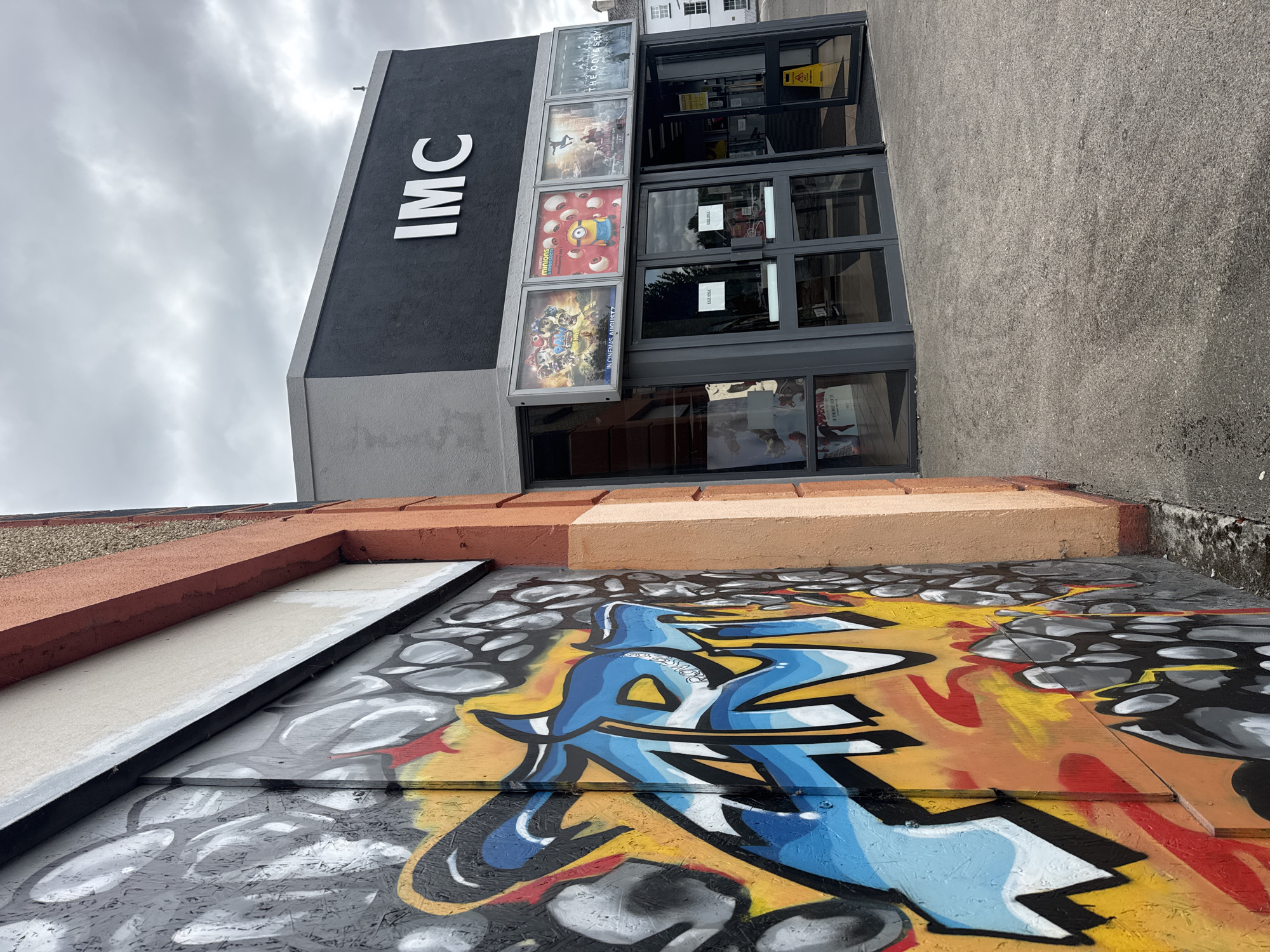This image captures the exterior of the IMC cinema in Clonmel, Ireland. The foreground is dominated by a brightly colored graffiti mural on the side of a building, featuring abstract shapes in blues, yellows, and reds against a backdrop of grey and black. To the right, the cinema building itself is visible, with its dark grey facade displaying the "IMC" logo in white, illuminated letters. Above the entrance, four movie posters are displayed in individual frames. The entrance consists of double glass doors, some of which are propped open, revealing the interior. A yellow "wet floor" sign is placed near the open entrance. The pavement in front of the cinema is a weathered, grey concrete. The sky above is overcast and cloudy, suggesting a temperate climate. The architectural style of the cinema appears modern. The overall scene is a juxtaposition of urban street art and commercial entertainment.