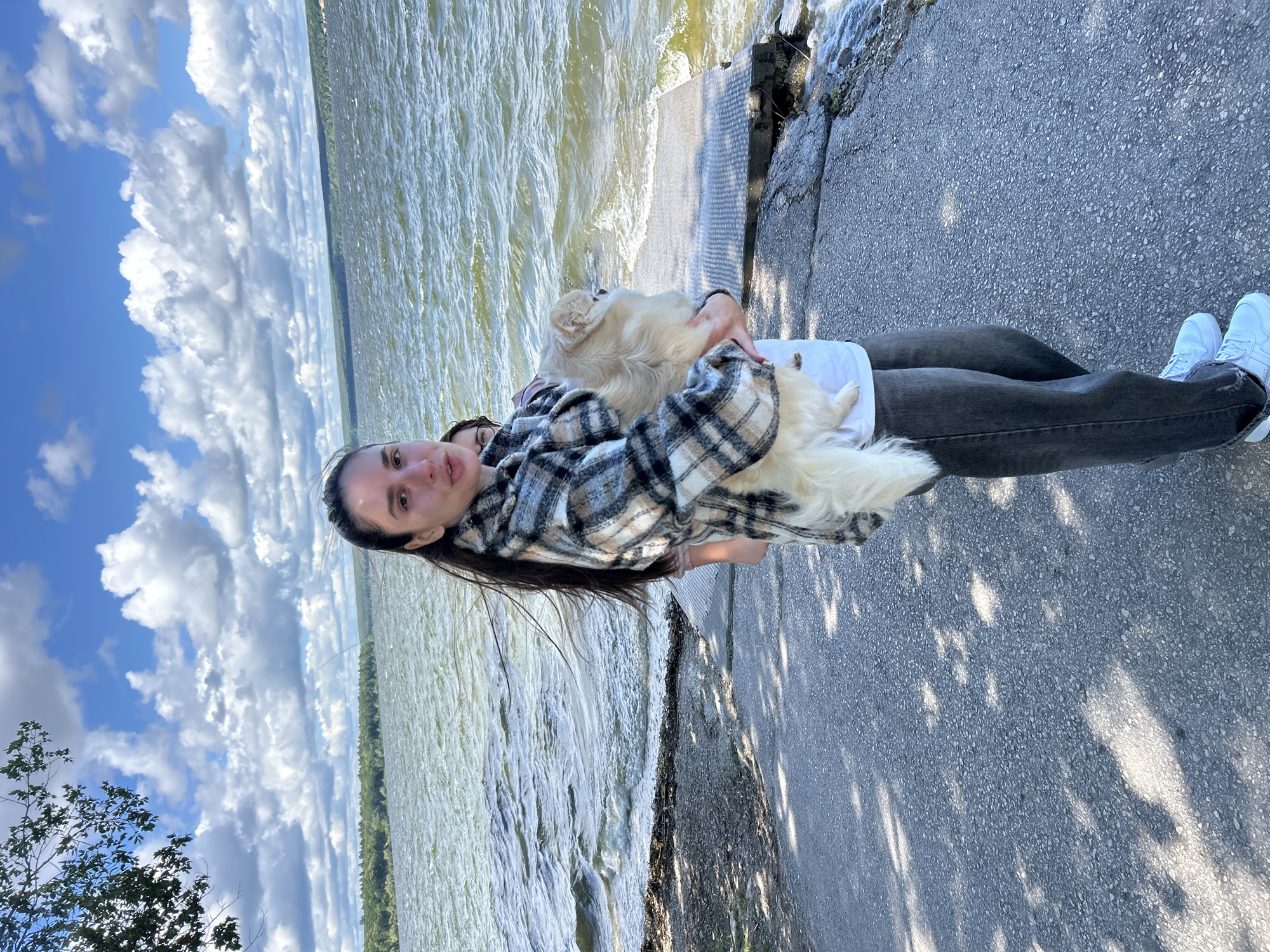A young woman with long, dark hair is holding a fluffy, light-colored dog. She is wearing a plaid jacket over a white shirt and dark jeans, with white sneakers. She is standing on a paved area near the water. In the background, there is a large body of water with choppy waves, and a tree-lined shore with a distant horizon under a blue sky with white clouds. A concrete structure, possibly a dock or pier, is partially visible in the water. The woman is looking towards the camera, while the dog is looking away. There is another person, partially obscured, behind the woman. The location is not specified as Междуреченск, Russia, and the scene appears to be a natural outdoor setting.