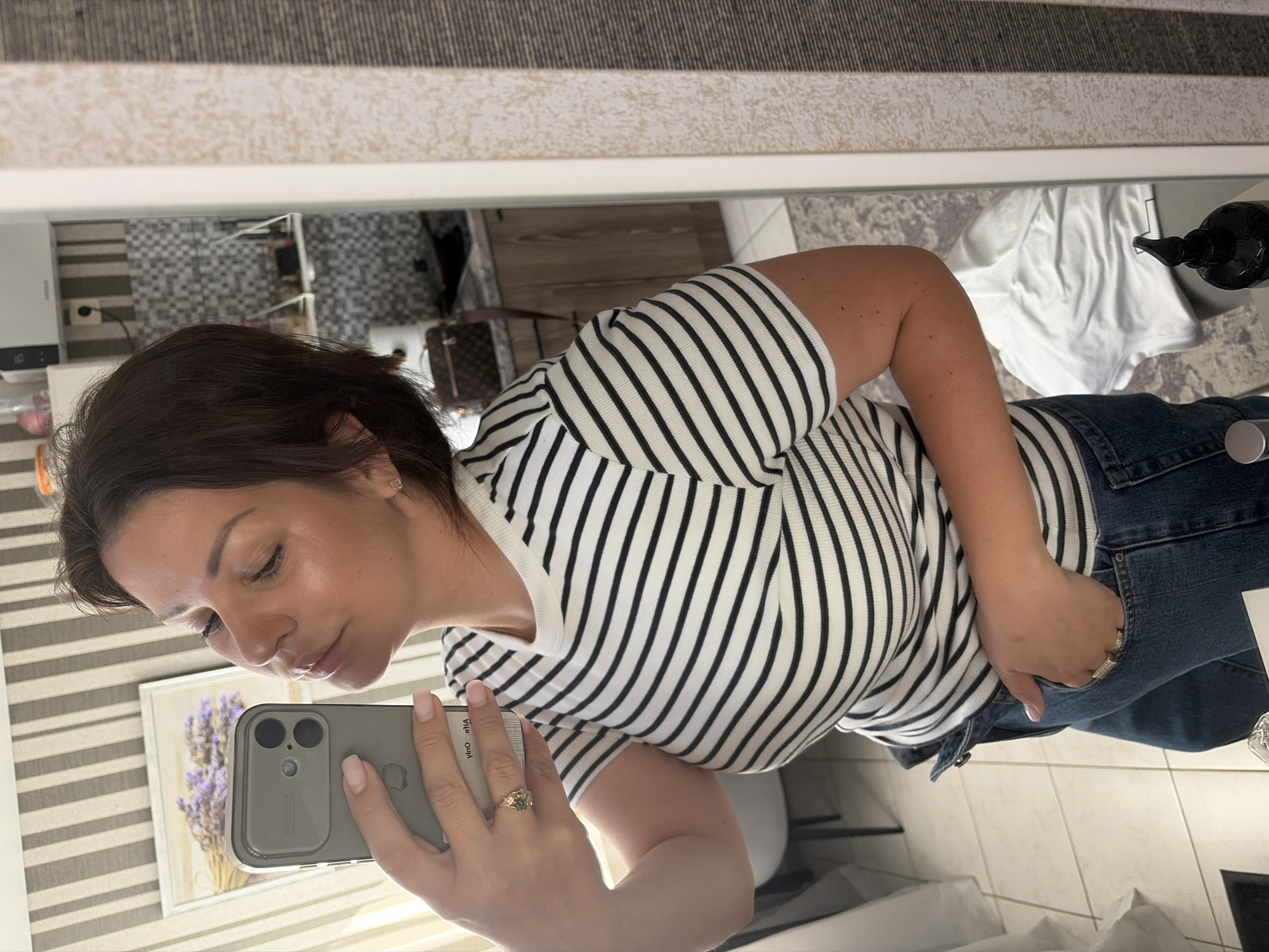 A woman is taking a selfie in a room. She is wearing a black and white striped t-shirt and blue jeans. She has her phone in her left hand and her right hand in her pocket. The background shows a striped wall, a framed picture, a mirror, and kitchen cabinets. The room appears to be a domestic interior.