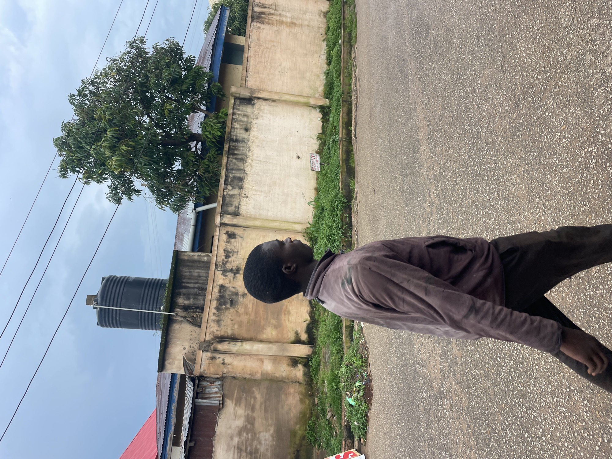 A young Black male is walking on a paved road, moving from left to right across the frame. He is wearing a dark brown long-sleeved shirt and dark pants. His posture suggests he is walking with purpose. The background features a weathered, light-colored concrete wall that extends horizontally across the image. Behind the wall, there are structures with corrugated metal roofs, some appearing aged and rusted. A large black water tank is visible on top of one of the structures, and a leafy green tree grows to the right of the tank. The sky is overcast, with a muted, cloudy appearance. The environment appears to be a residential or commercial area in Jos, Nigeria, characterized by older buildings and infrastructure.