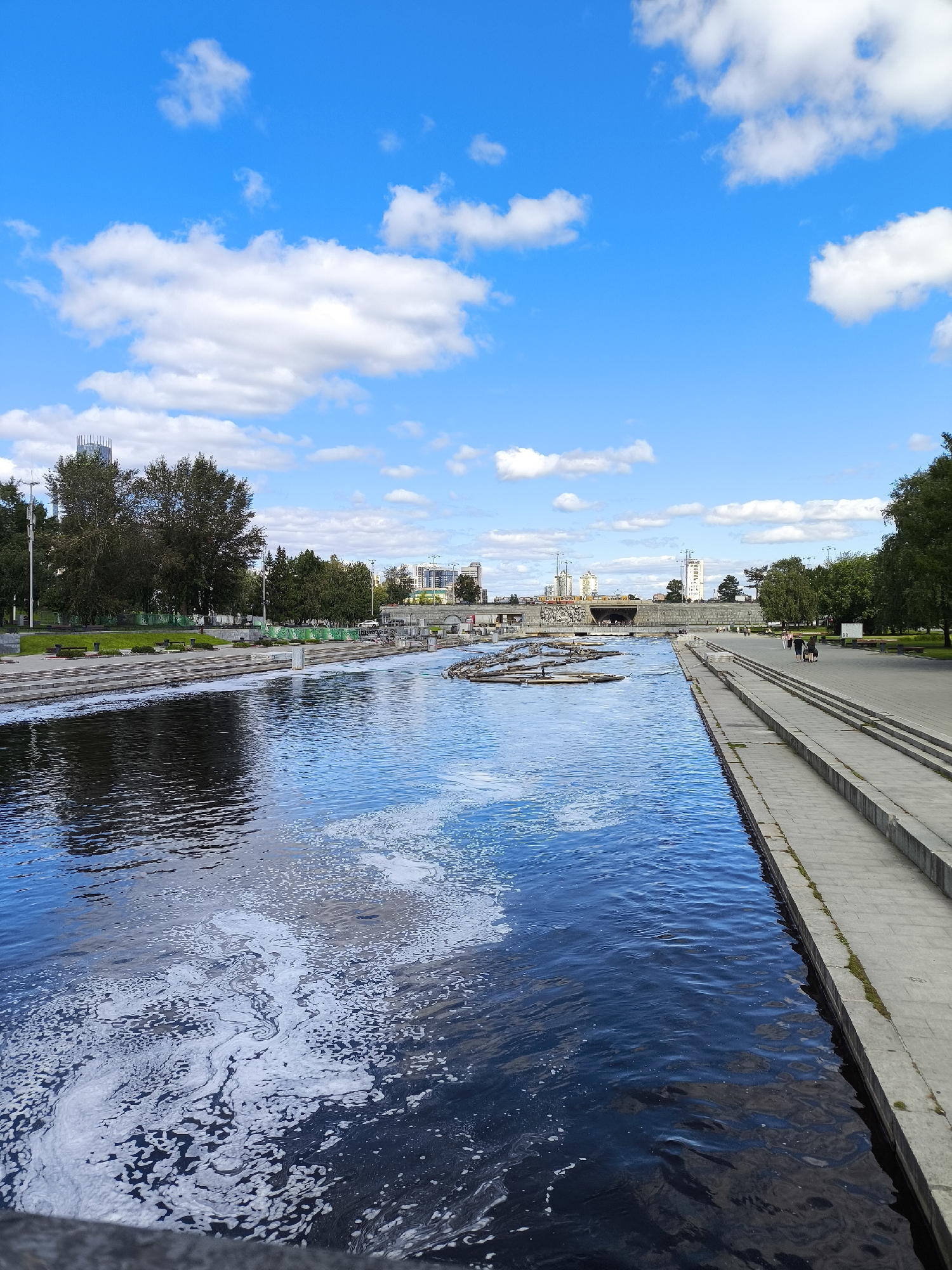 The image captures a view of the Iset River in Yekaterinburg, Russia, on a sunny day with scattered white clouds against a vibrant blue sky. The river occupies the foreground, its surface disturbed by ripples and patches of white foam. The left bank of the river is lined with trees and green grass. Across the river, a concrete embankment with tiered steps leads up to a walkway where several pedestrians are visible, some walking in pairs and others individually. Beyond the embankment, a cityscape with modern buildings and construction cranes can be seen. In the distance, a bridge spans the river. The overall scene depicts a serene urban waterfront with a mix of natural elements and architectural structures.
